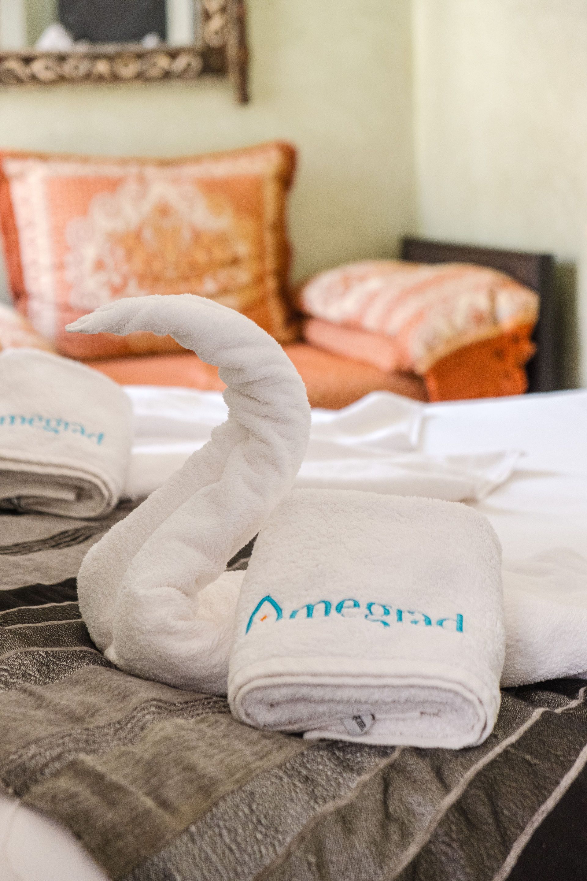 White towel swan on bed, with folded towels, and decorative pillows in the background.