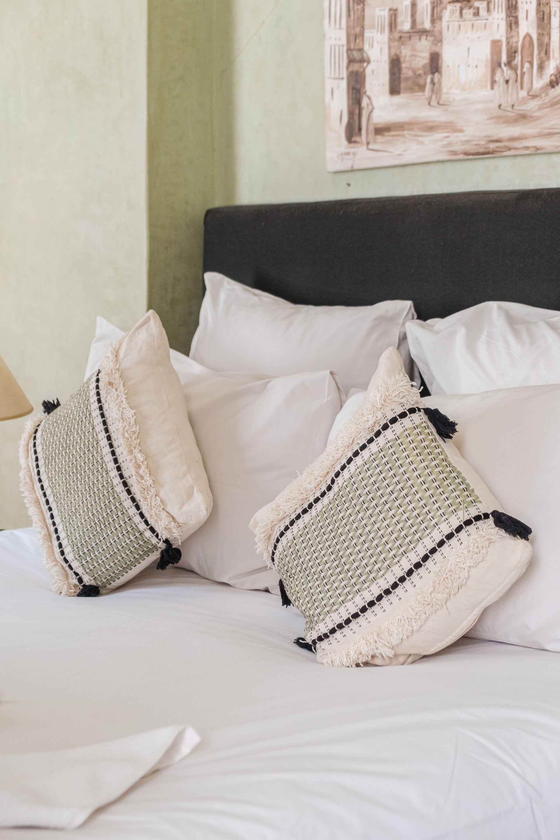 Bed with white bedding, two patterned pillows, against a green wall with a framed painting.