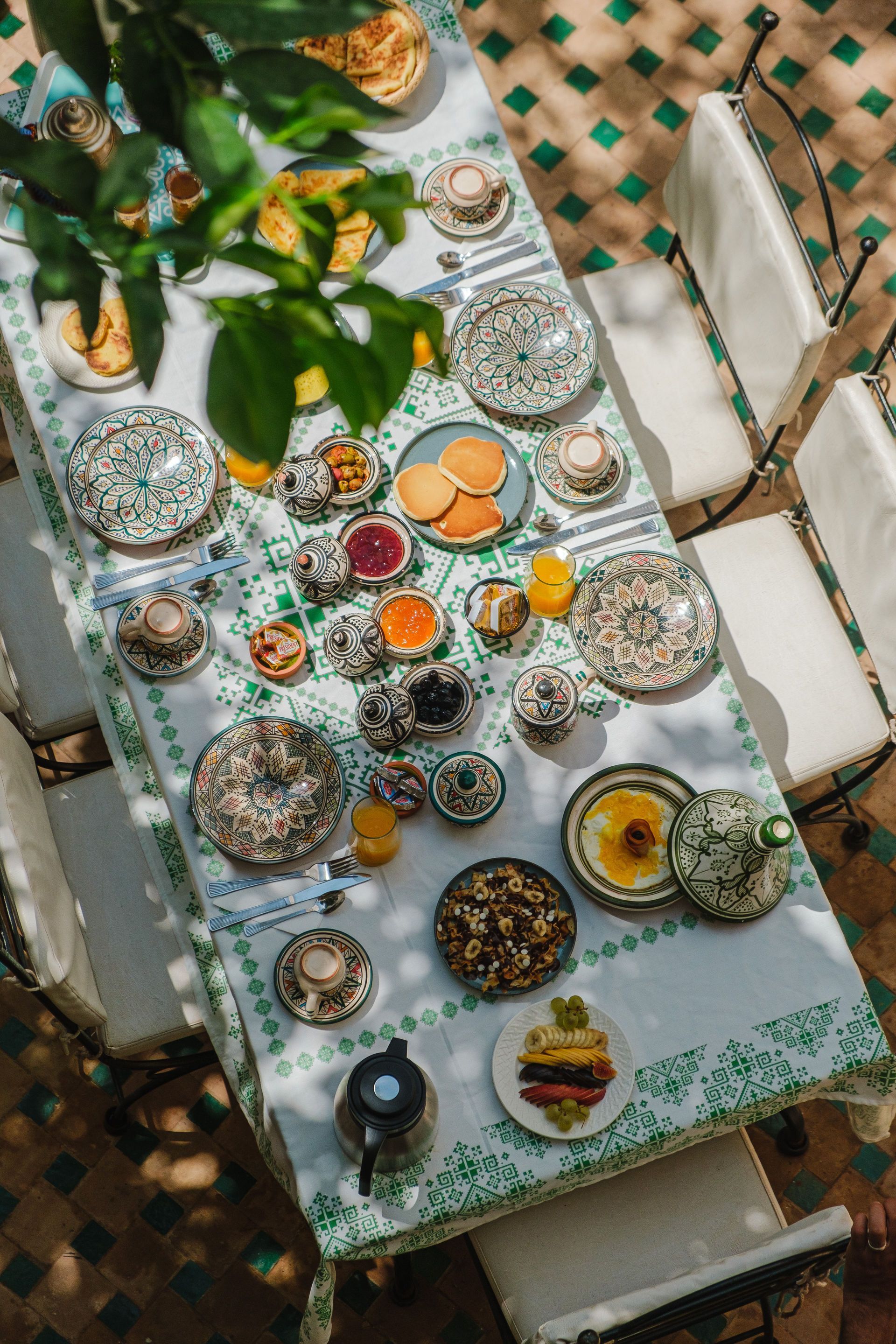 A table set for brunch outdoors with patterned plates, food, and drinks. Sunny, green tiled floor.