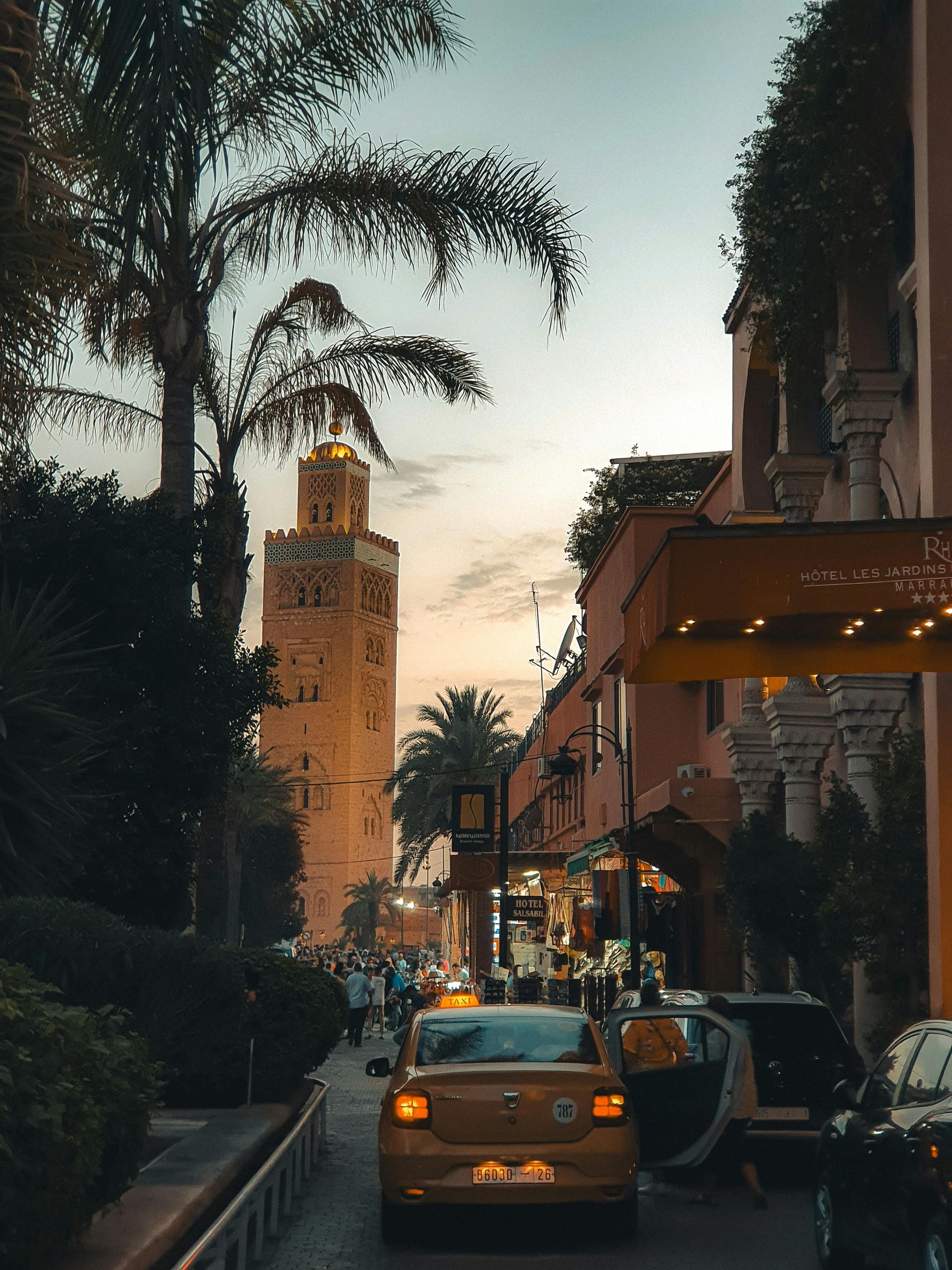 A tall mosque minaret in Marrakech, Morocco, next to a busy street with cars and a hotel at dusk.