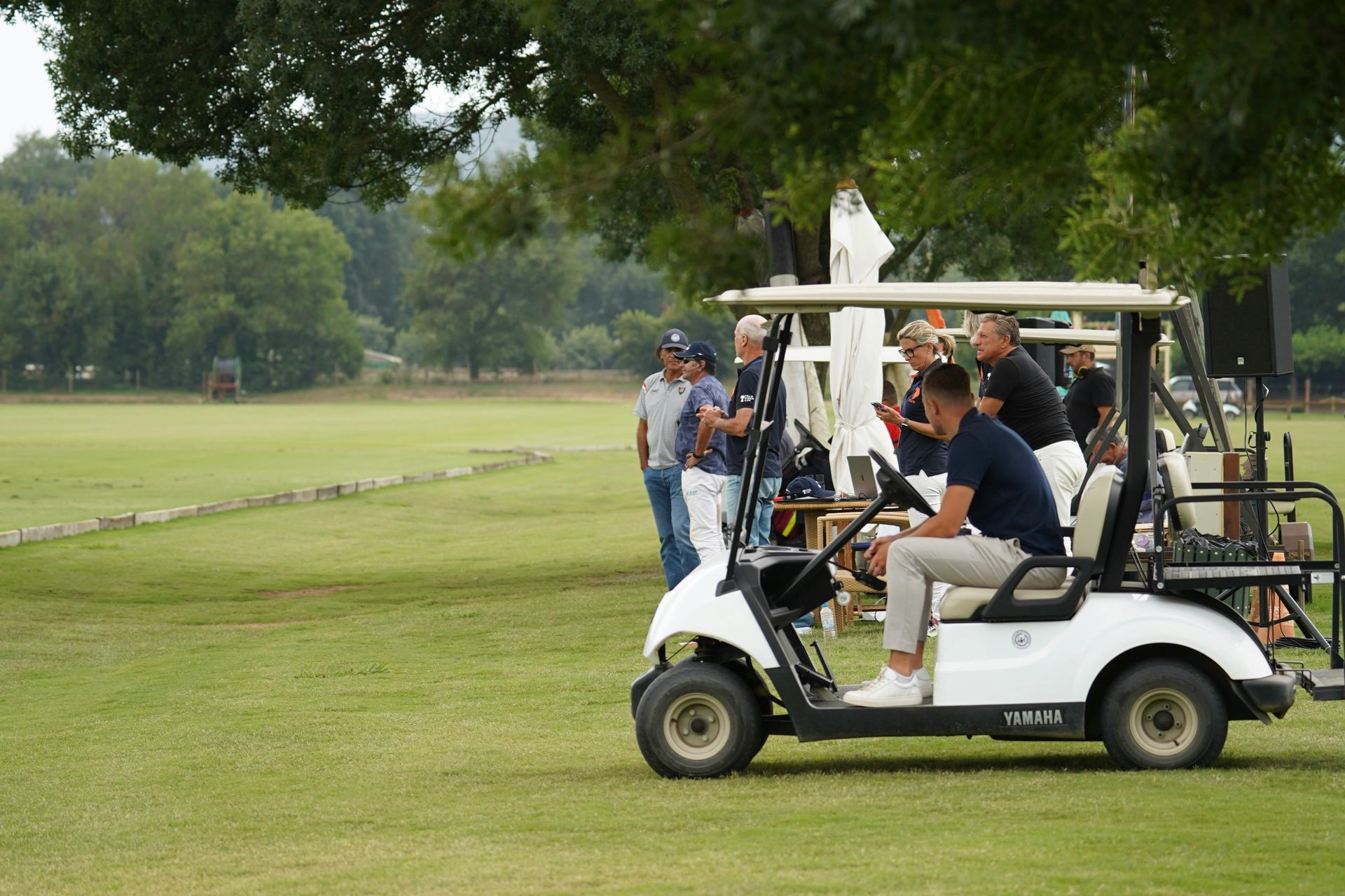 A group of people standing by and sitting in a white golf cart on a grassy field at an outdoor sporting event.