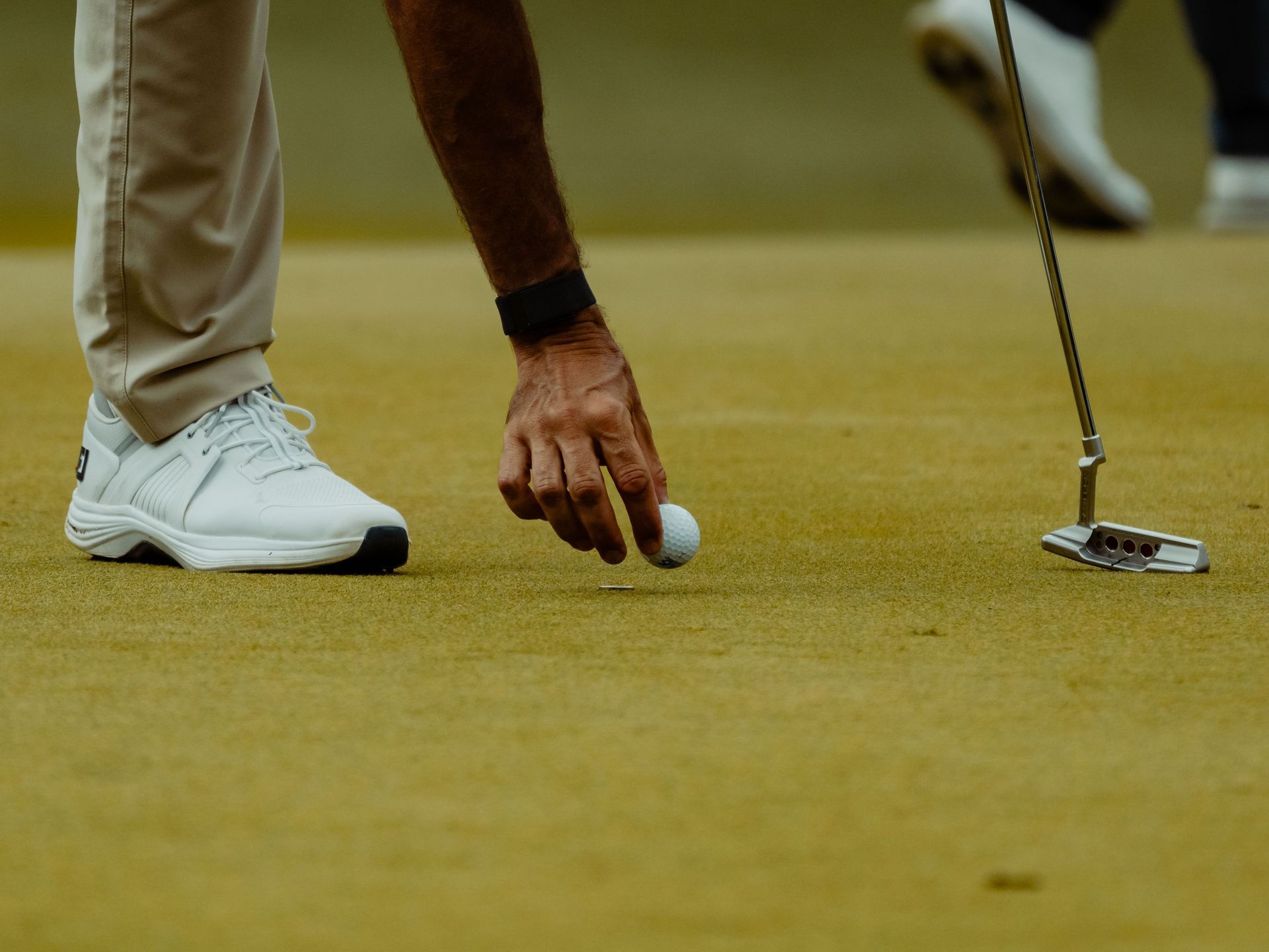 Golfer placing a white ball on a green putting surface beside a putter and shoes