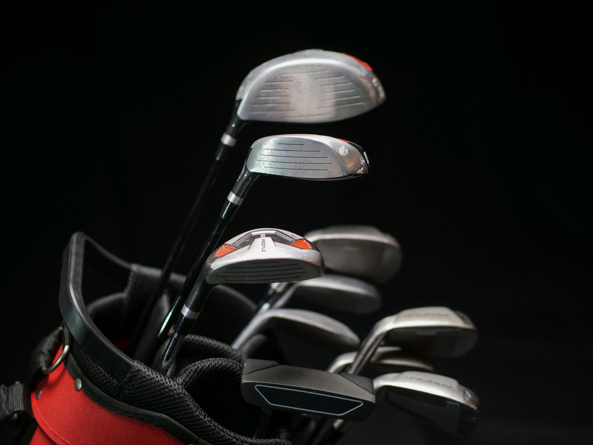 A set of golf clubs with silver heads and black shafts standing upright in a red and black golf bag against a black backdrop.