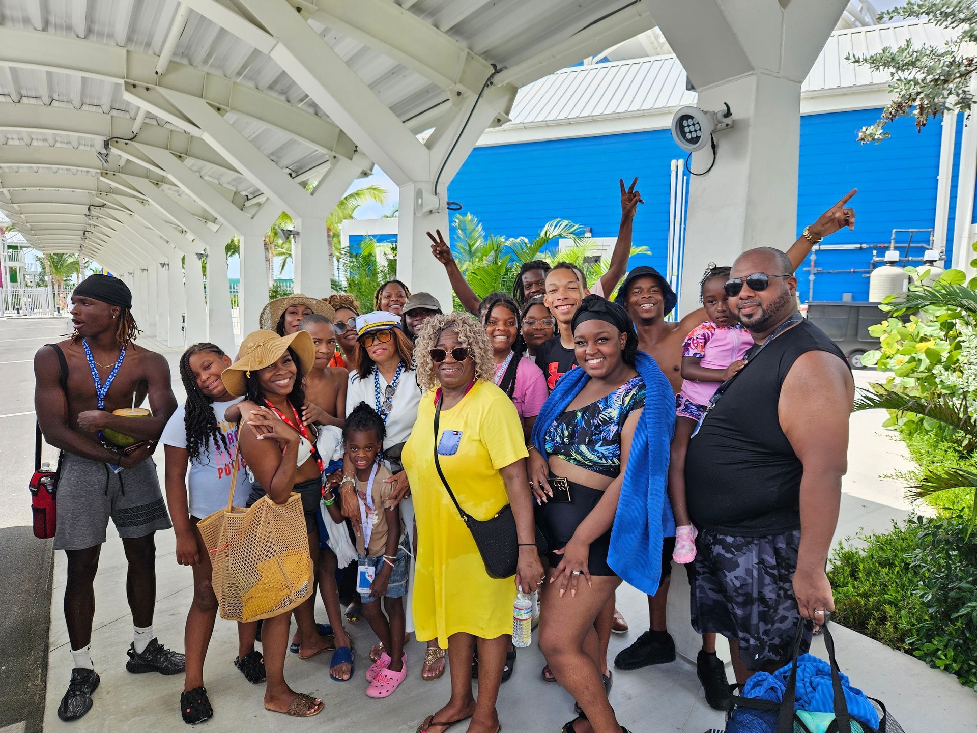 Group of people posing outdoors under a white roof. Some smile, wave, and point at a blue building.