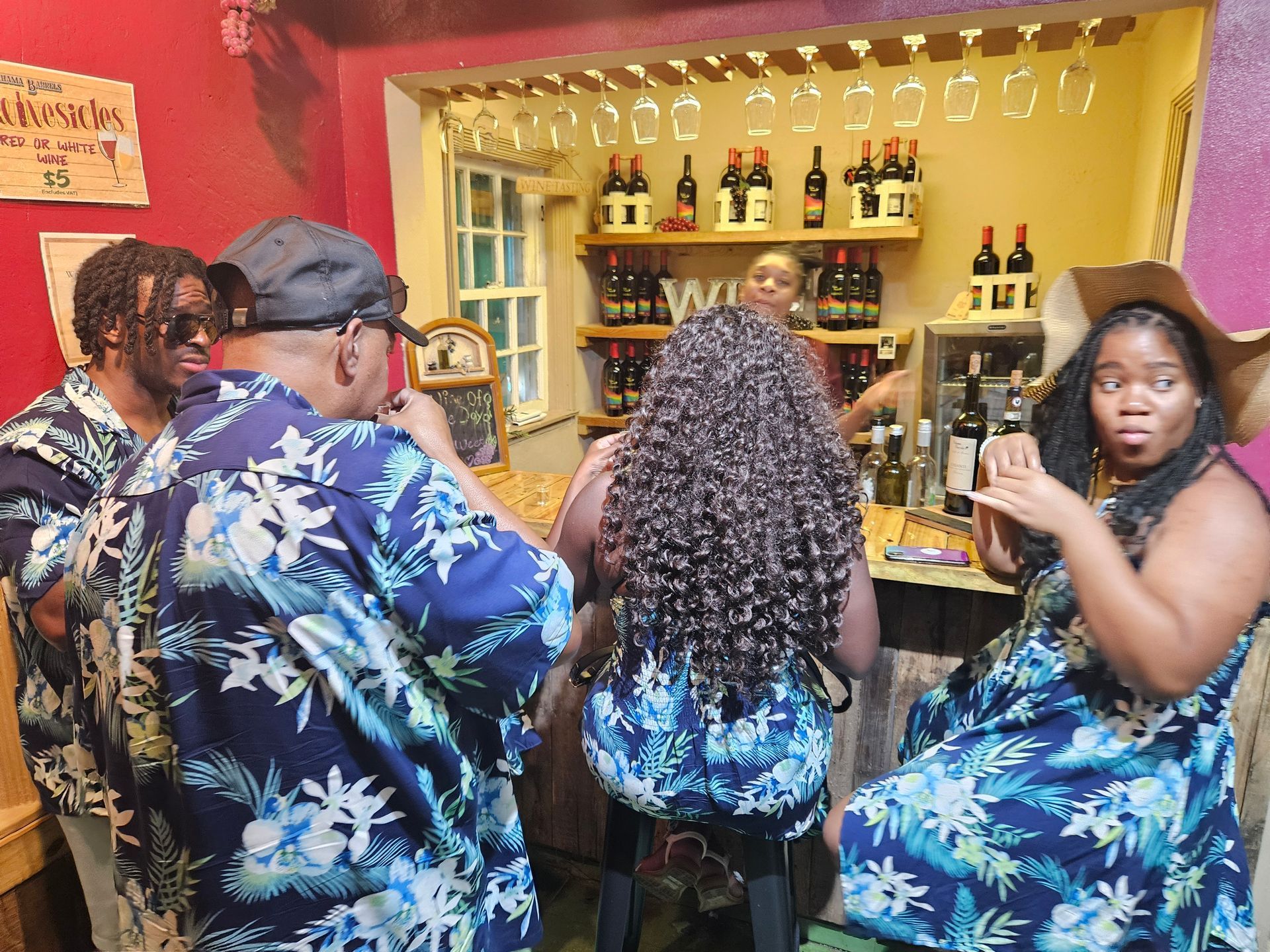 People at a wine bar; one woman looks surprised, others wear tropical print. Wine bottles and glasses visible.