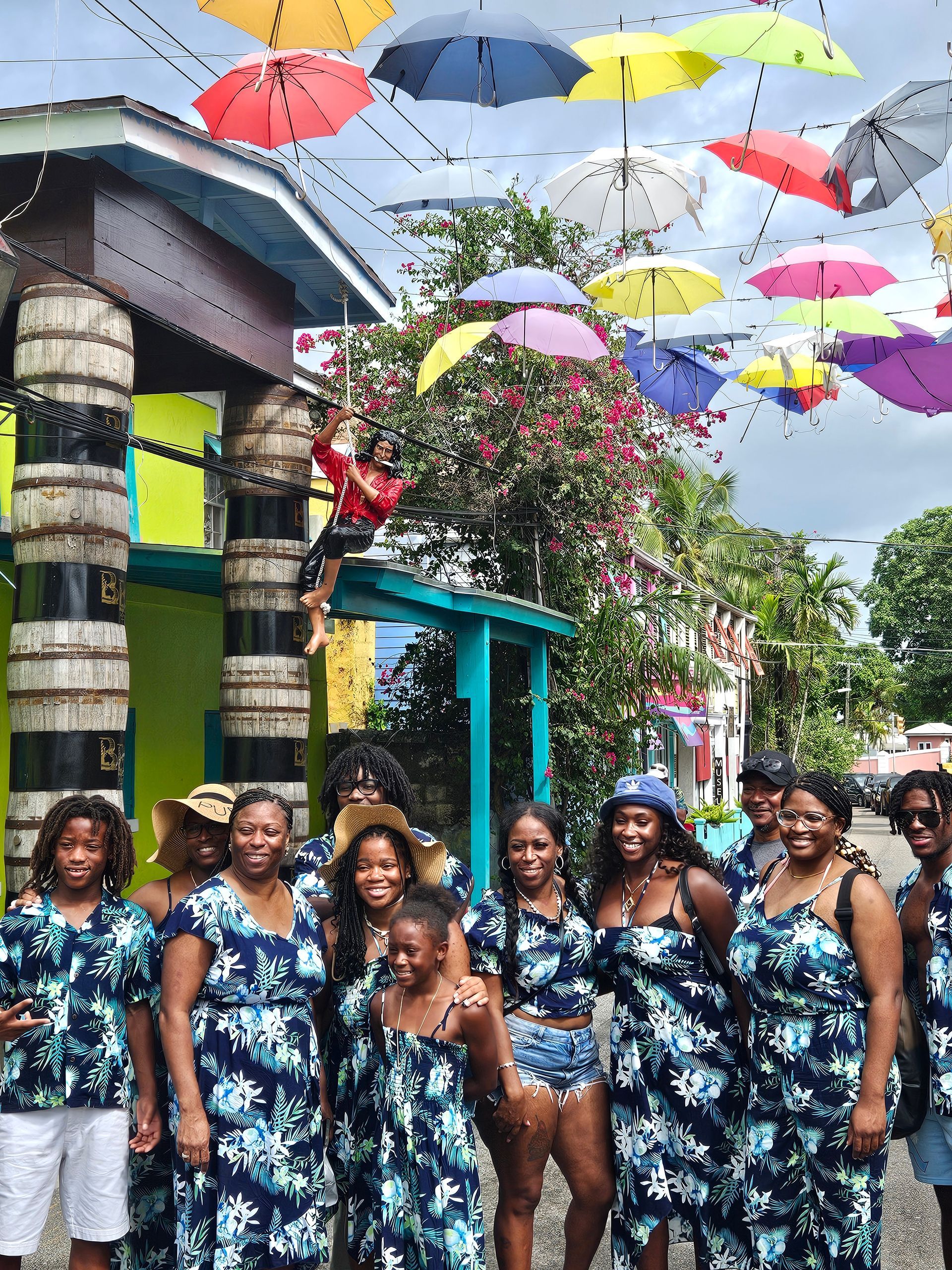 Group of people posing, wearing matching floral outfits, under colorful umbrellas strung above.