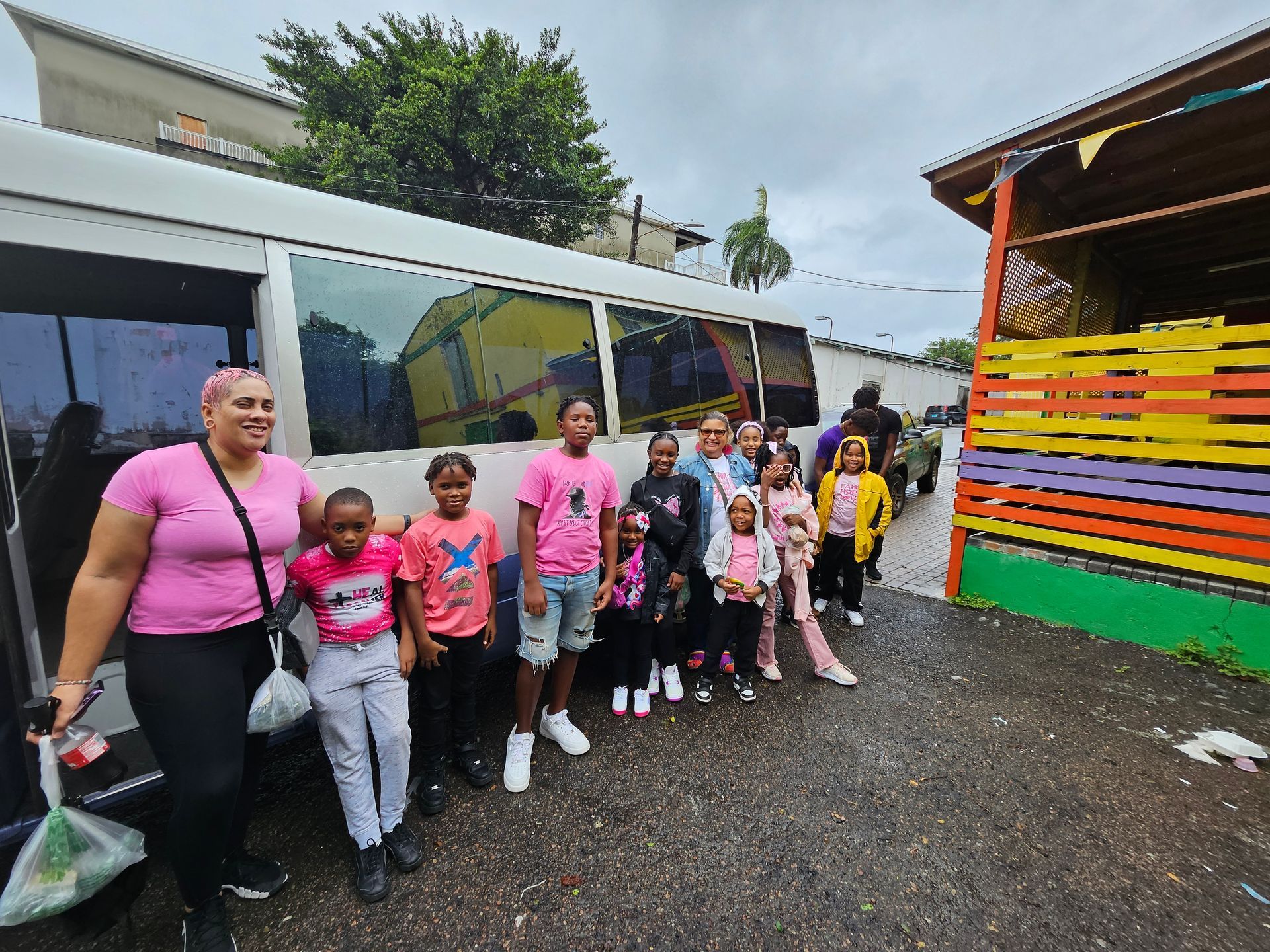 Group of children and an adult stand beside a white bus, likely about to board. Colorful building in background.