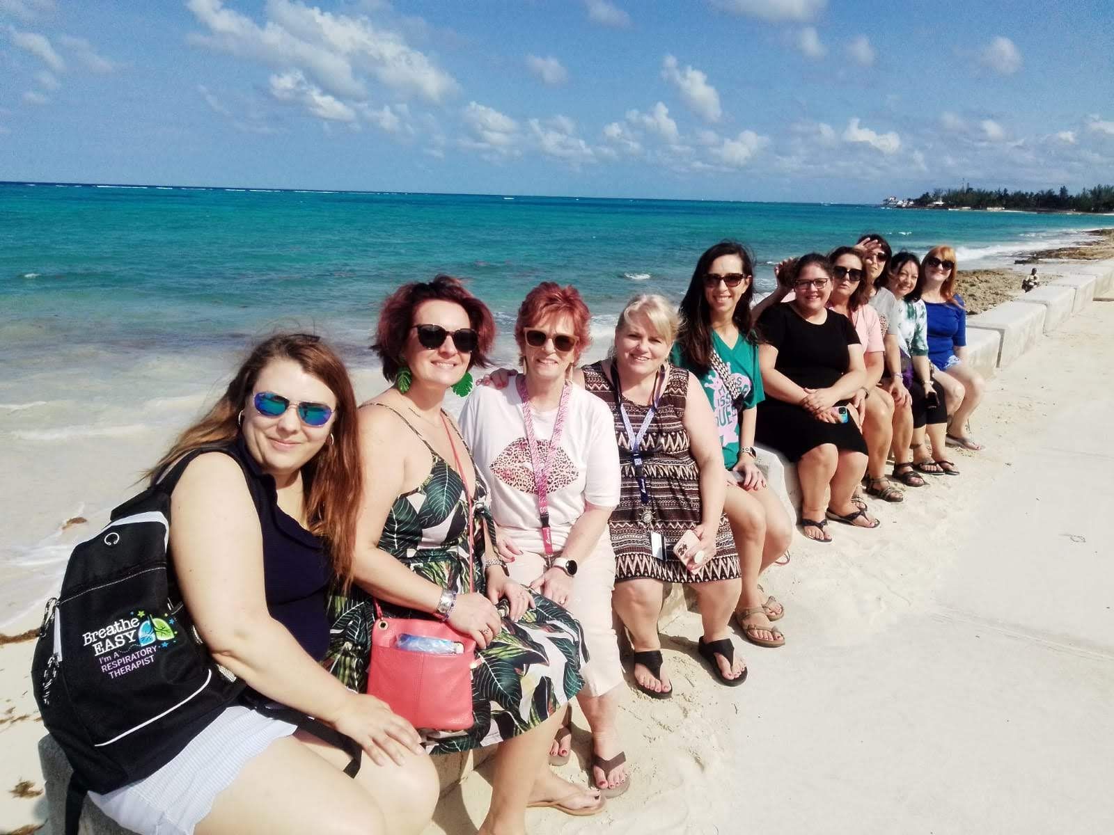 Group of women sitting on a sea wall, posing on a beach. Turquoise water, blue sky.
