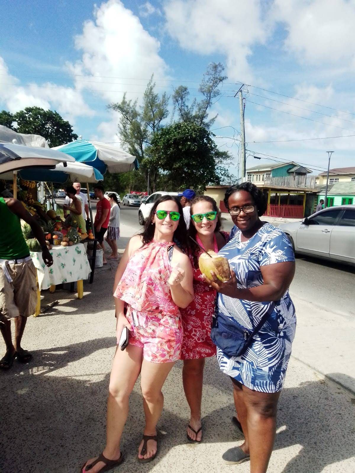 Three women smile outside, one holding a coconut. They wear sunglasses and summer clothes, with market stalls behind them.