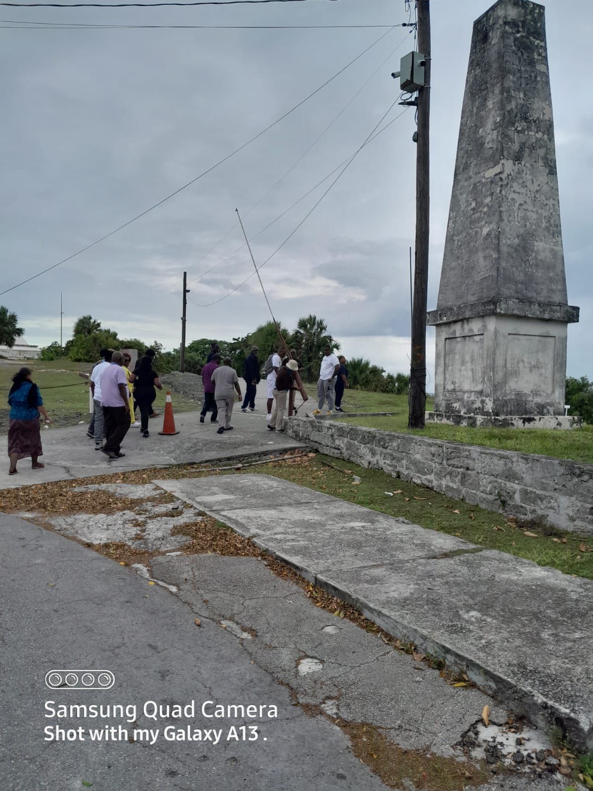 People walking near a tall, weathered obelisk on a cloudy day.