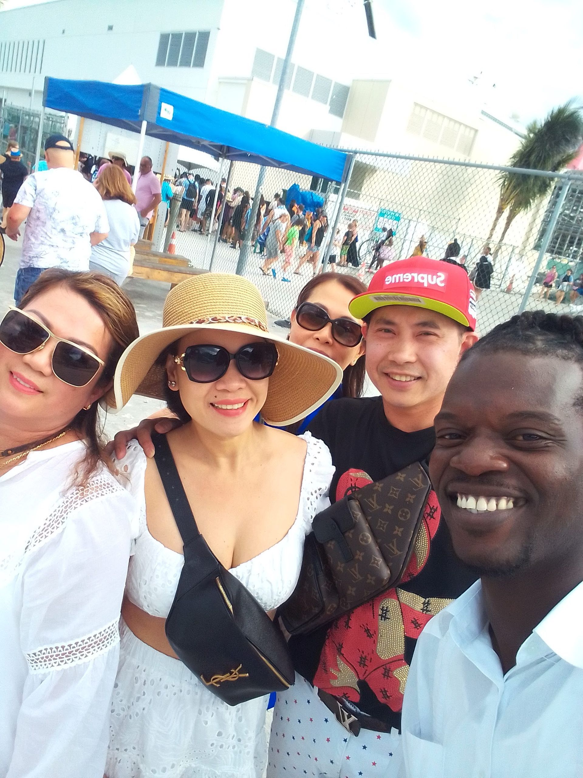 Group of people smiling for a photo outdoors near a fence, with a crowd and blue awning in the background.