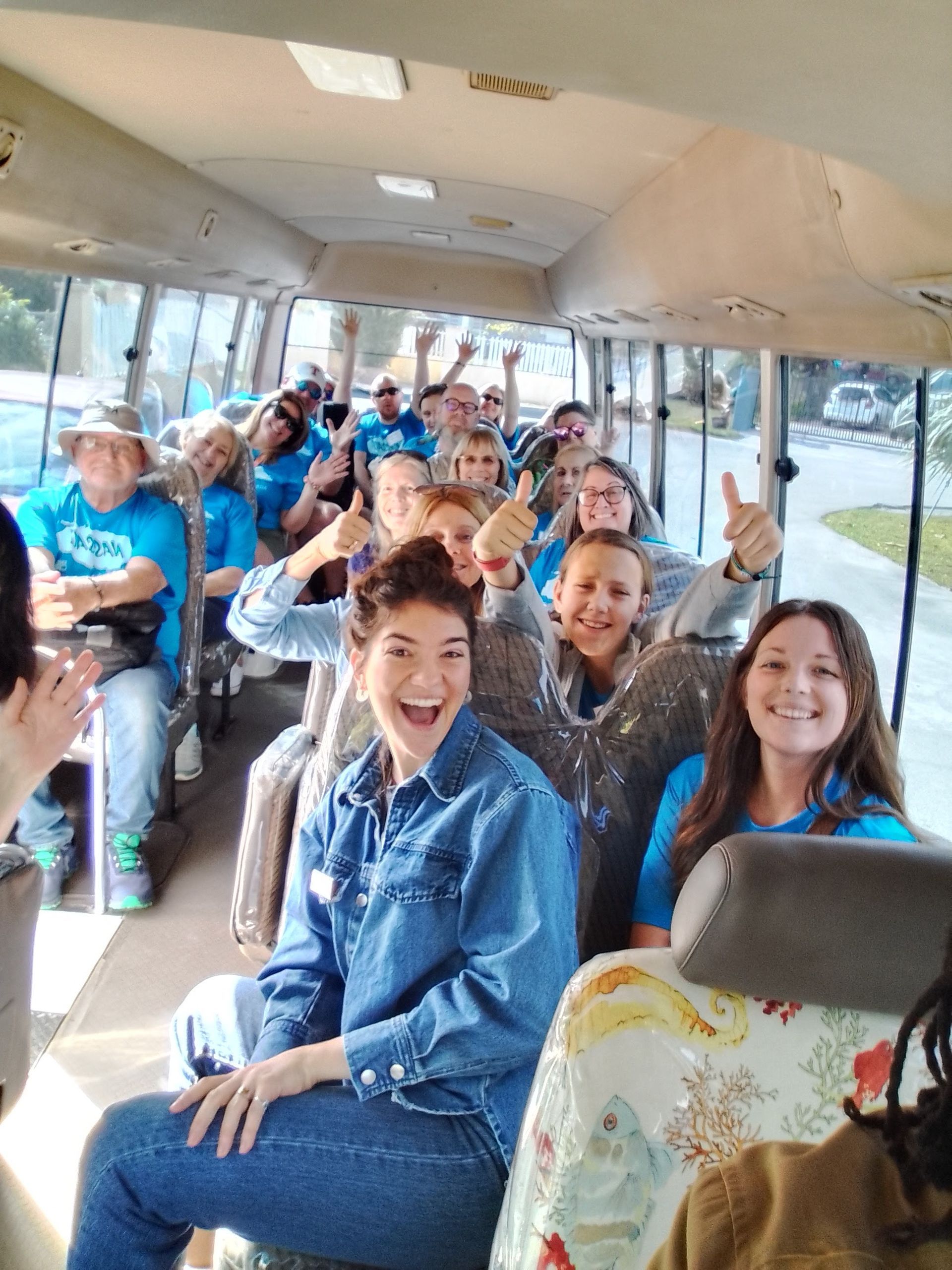 People inside a bus, many in blue shirts, smiling and giving thumbs up.