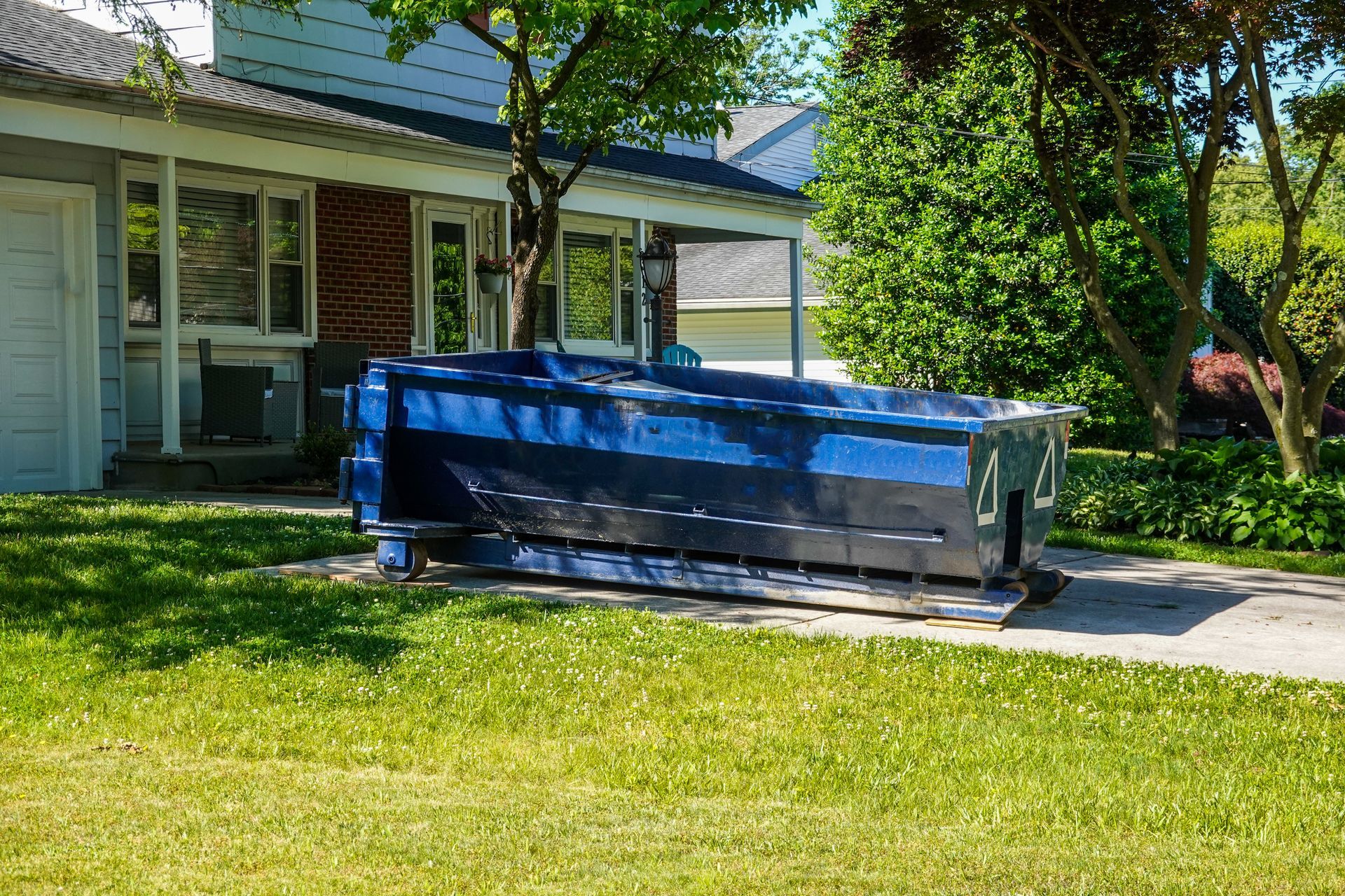 A blue dumpster is sitting in the grass in front of a house.