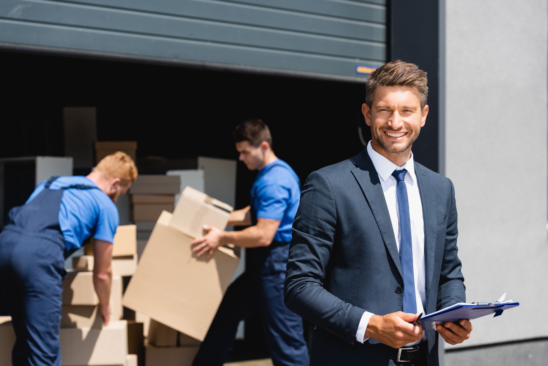 A man in a suit and tie is holding a clipboard in front of a warehouse.