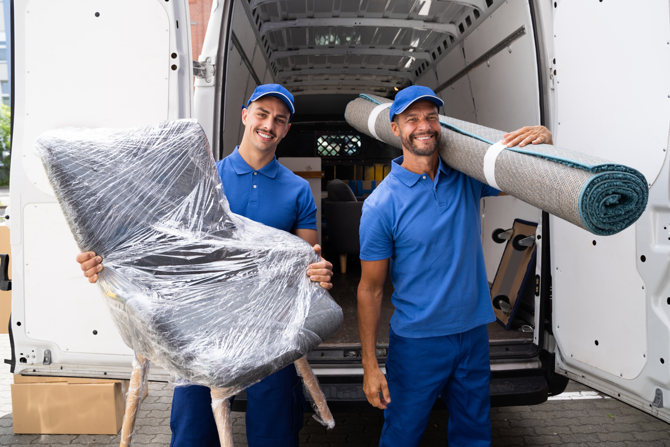 Two movers are carrying a chair and a rug in front of a van.