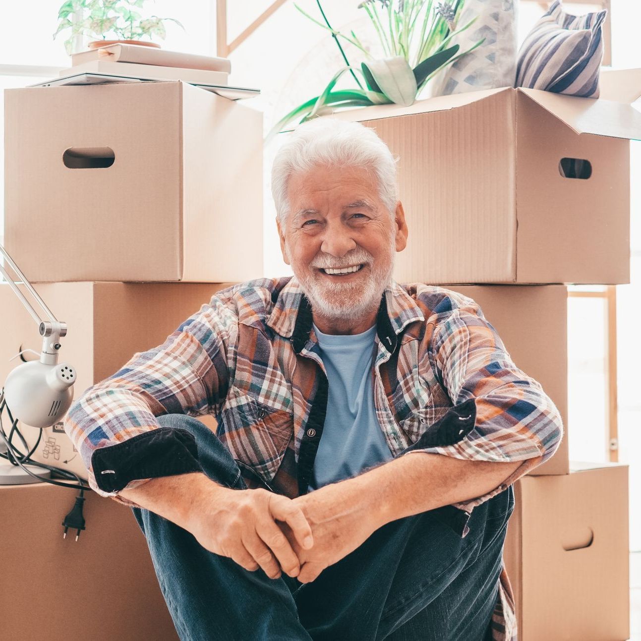 An elderly man is sitting on the floor in front of a pile of cardboard boxes.