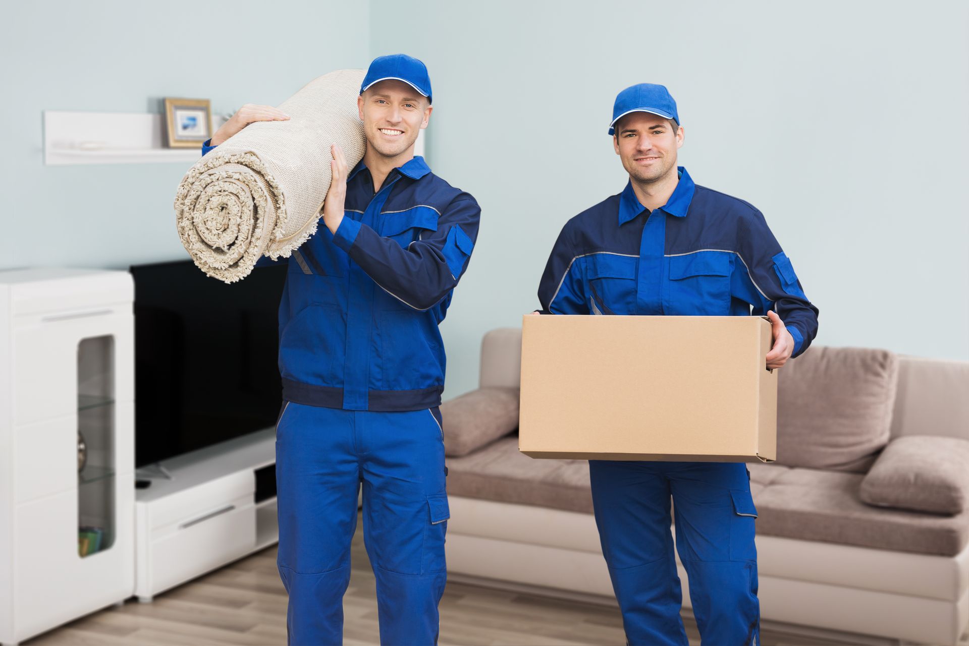 Two movers are carrying a rug and a box in a living room.