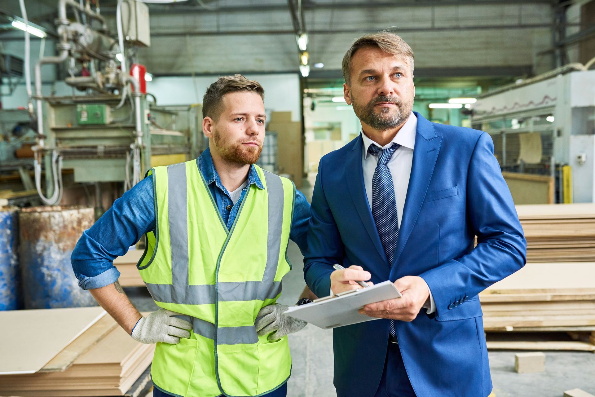 A man in a suit and tie is standing next to a man in a safety vest in a factory.