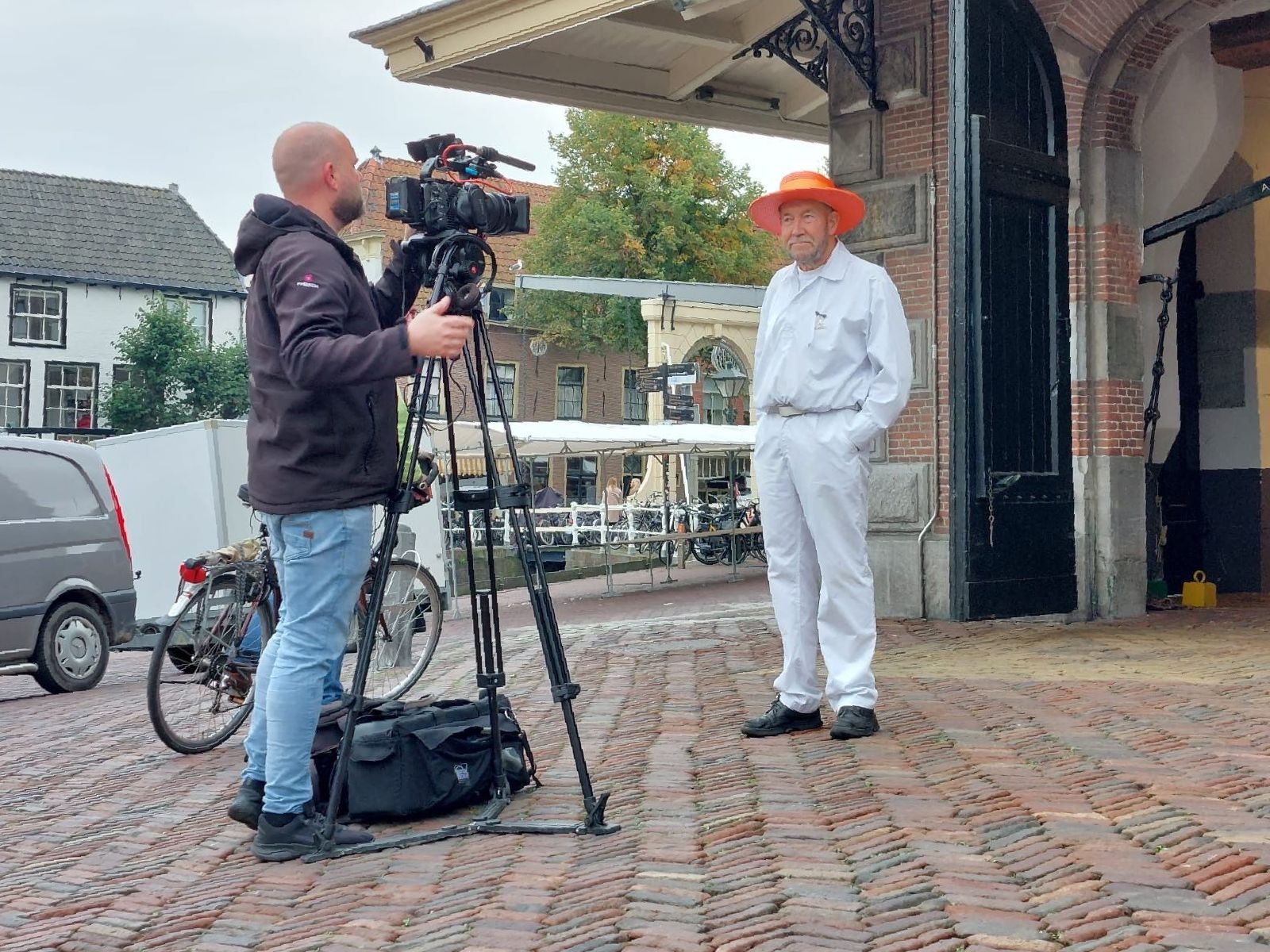 Een man in een wit pak en een oranje hoed wordt buiten geïnterviewd. Een cameraman filmt hem.