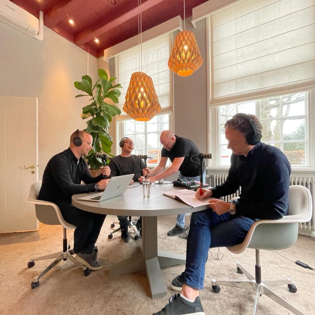 Vier mannen zitten rond een tafel in een lichte kamer en nemen een podcast op. Een van hen maakt aantekeningen.