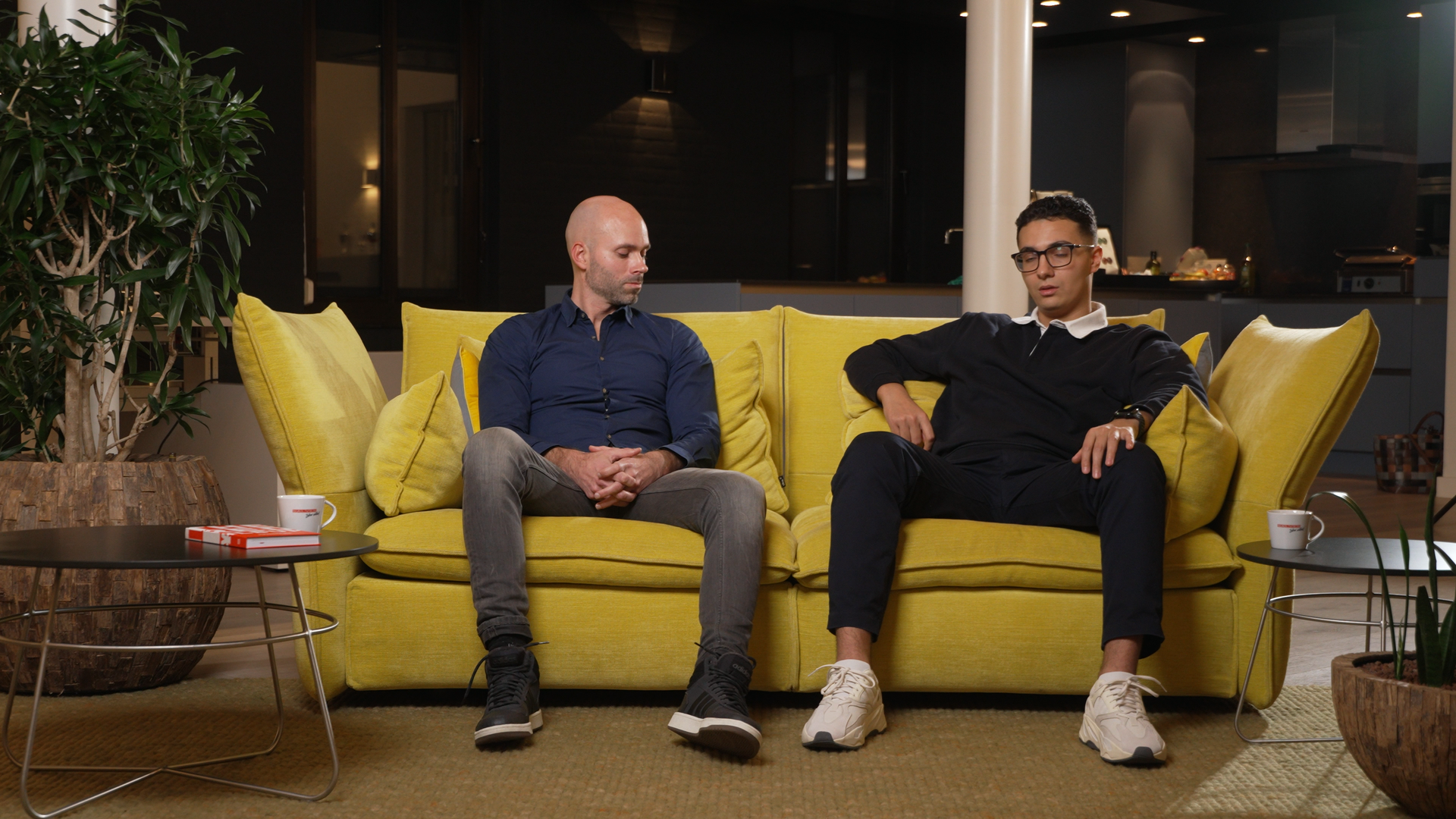 Twee mannen zitten op een gele bank in een moderne setting, de een in een blauw overhemd, de ander in een trui.