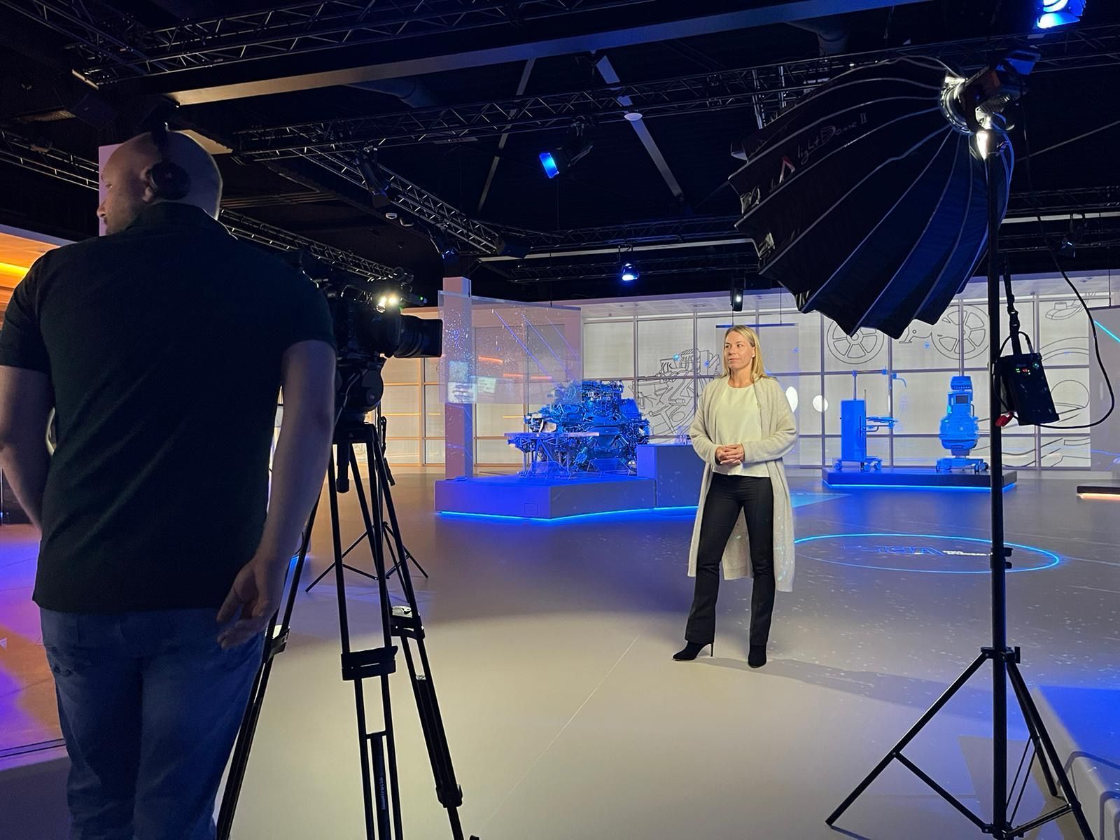 Een vrouw wordt gefilmd in een studio; een cameraman, camera, lichten en een decor met blauwe accenten.