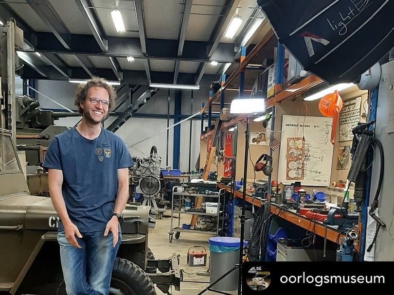 Man in garage glimlacht. Hij staat vlakbij een jeep, gereedschap en werkbanken. Goed verlichte werkplaats.