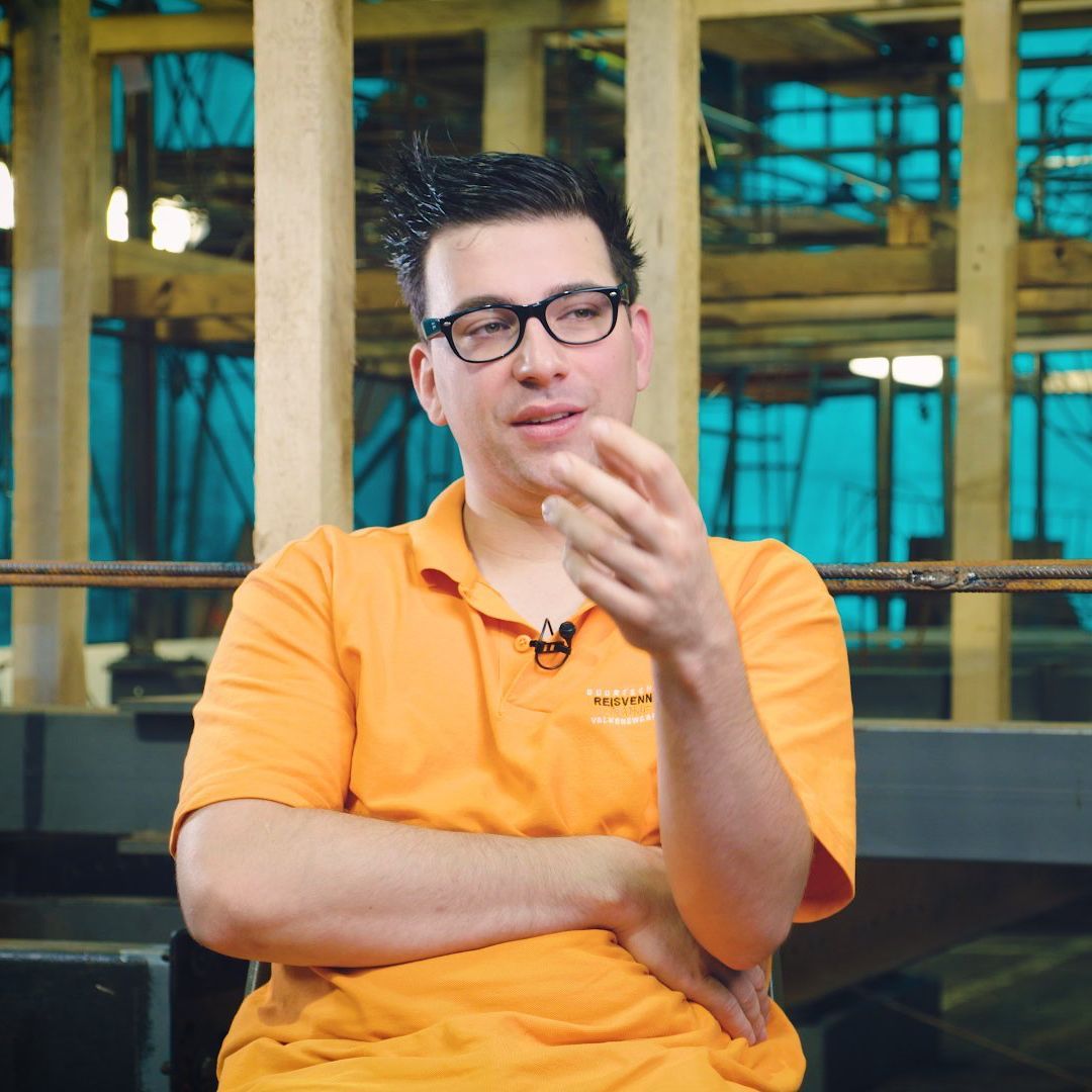 Man in oranje shirt en bril maakt een handgebaar; bouwsituatie.