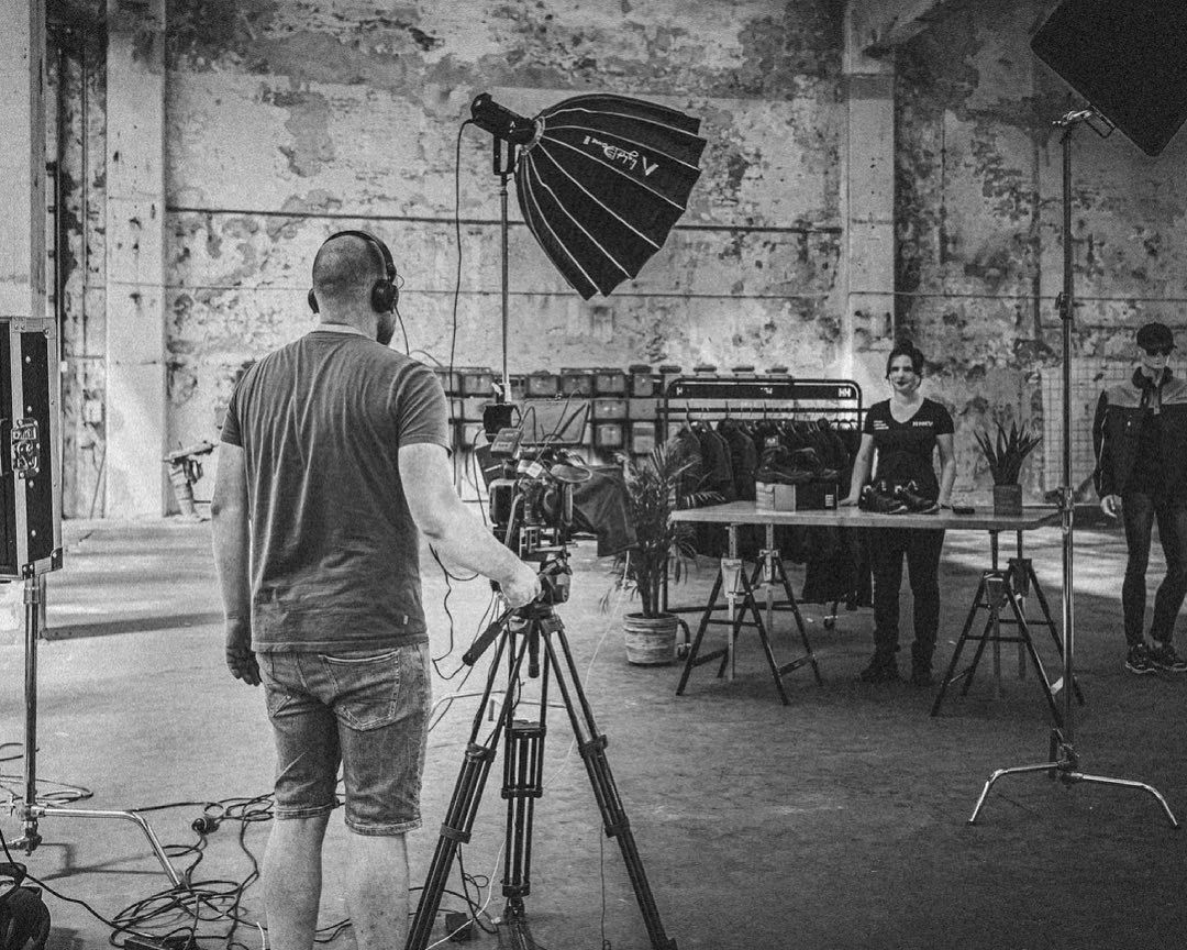 Een scène in een pakhuis wordt gefilmd. Een man bedient een camera op een statief; anderen staan ​​rond een tafel.