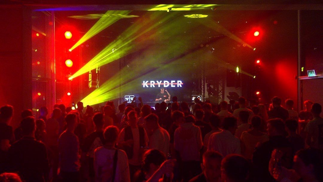 Publiek bij een concert met podiumverlichting, artiest KAYDER op het podium, rode en gele verlichting.