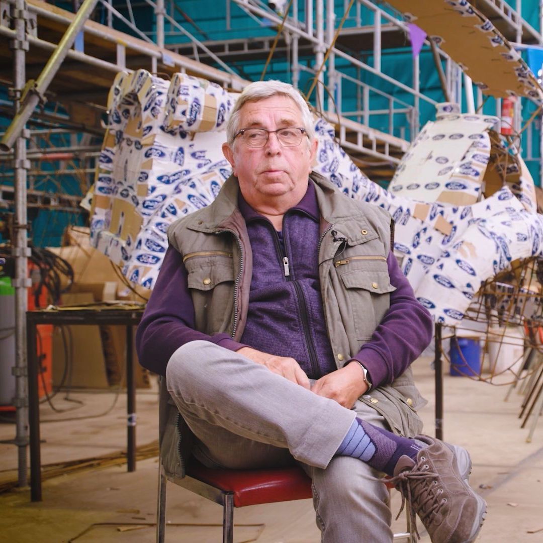 Man met bril zit voor een bouwwerk, gekleed in vest en paars shirt.