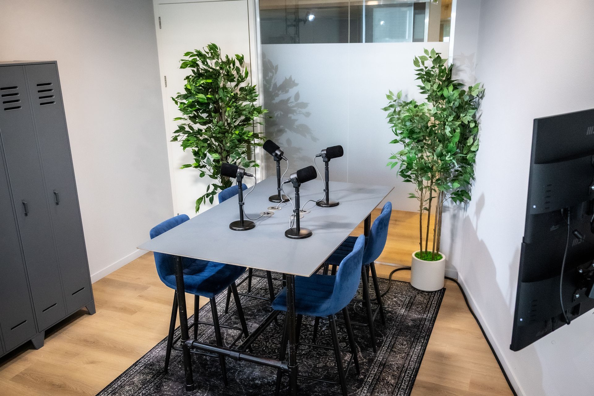Podcaststudio met microfoons op een tafel, blauwe stoelen, planten en een grijs kluisje.