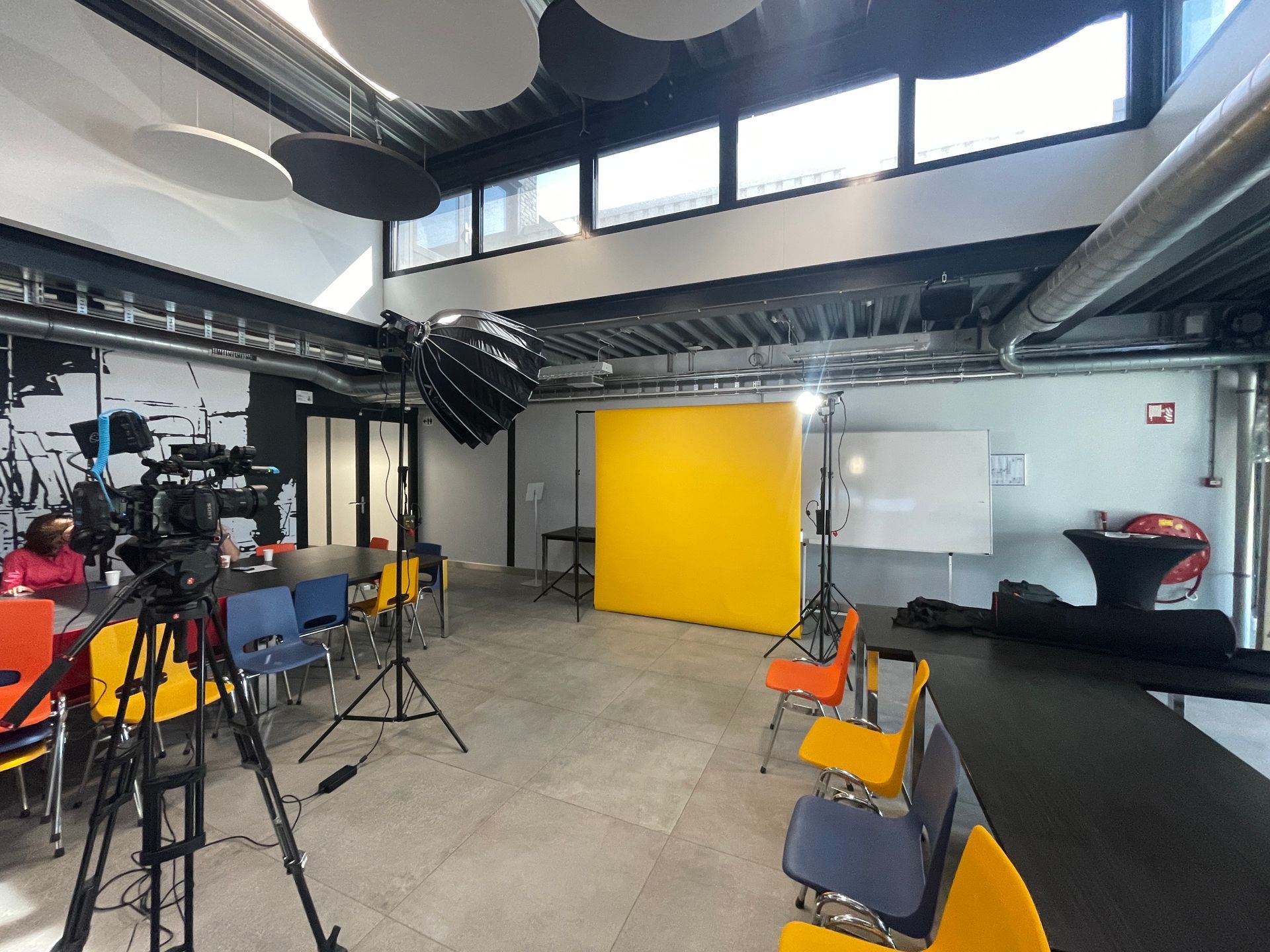 Een studio-opstelling met een gele achtergrond, camera's, verlichting, stoelen en tafels in een moderne kamer.