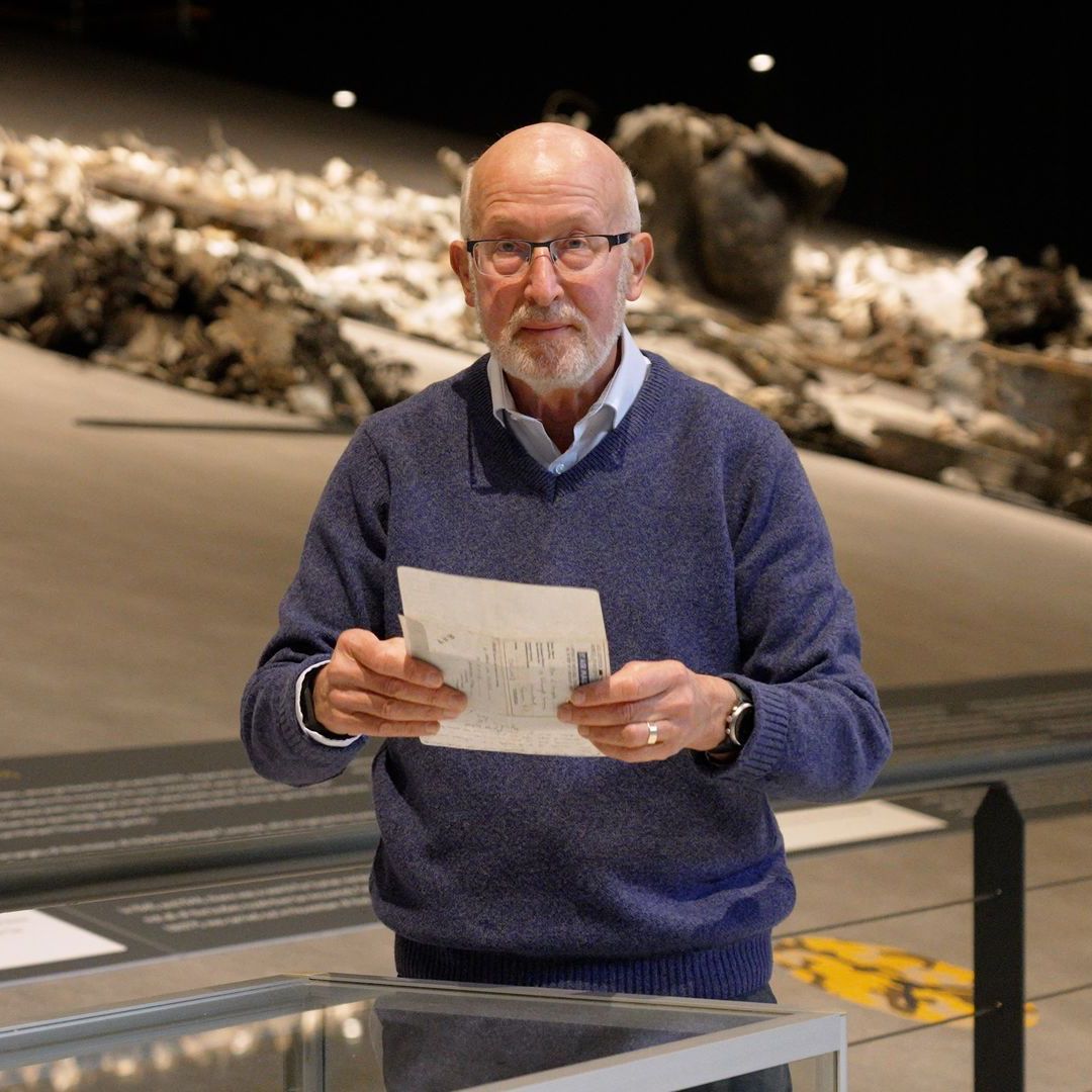 Man met bril in blauwe trui, papier vasthoudend, in museumtentoonstelling.