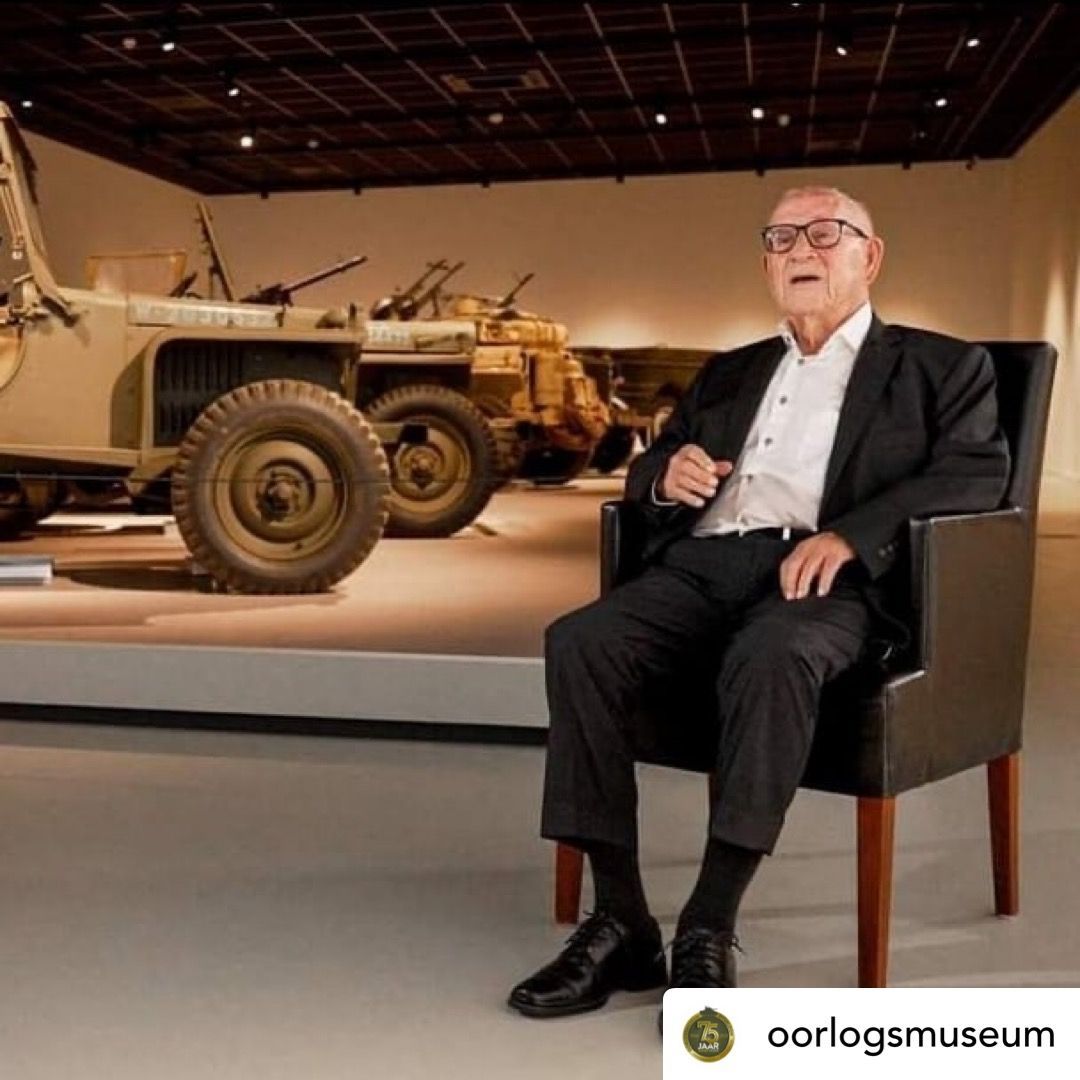 Man in pak zit op museumstoel; militaire voertuigen op de achtergrond.