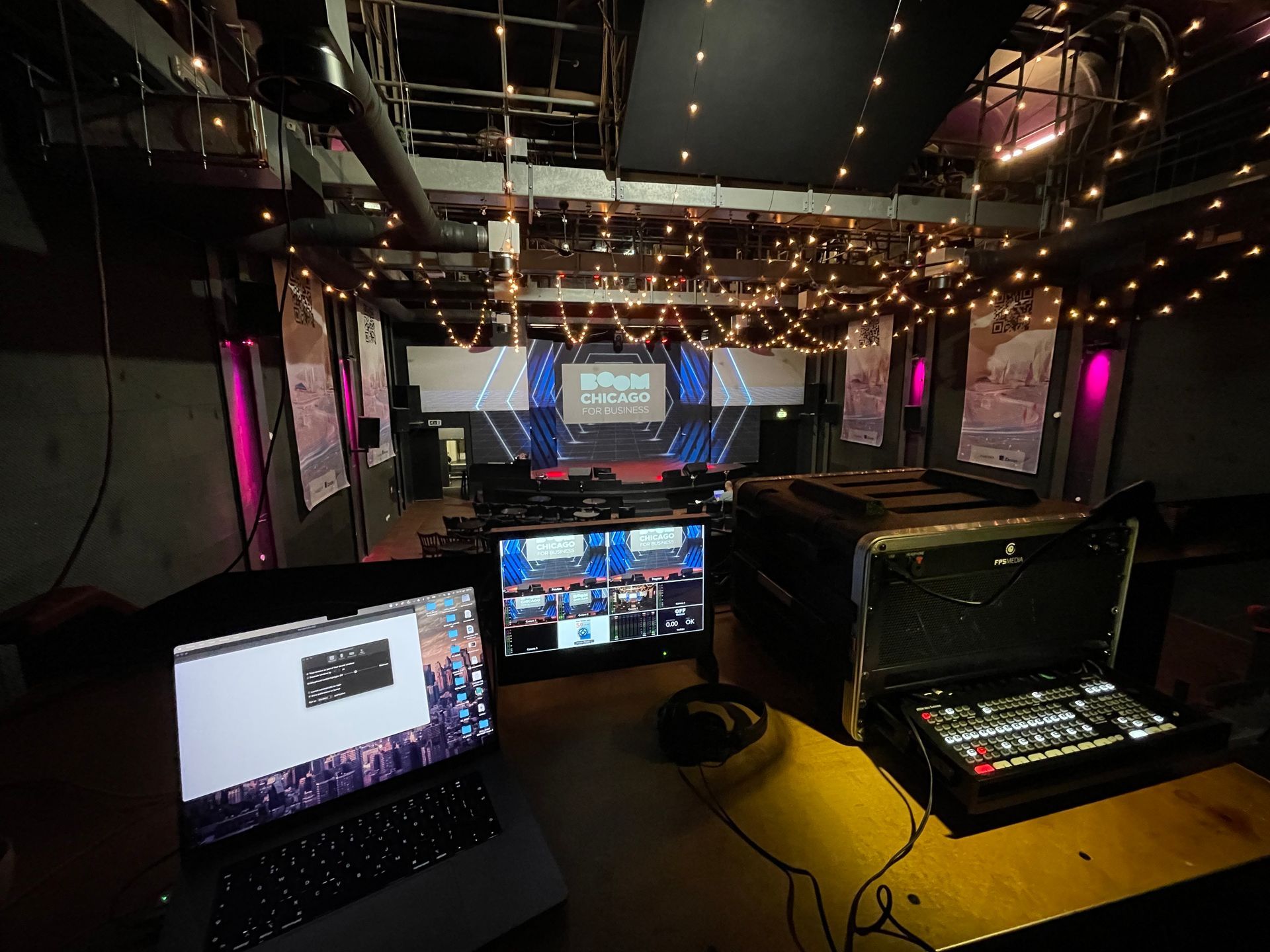 Laptop en beeldschermen op een tafel in een slecht verlichte evenementenruimte met een met licht versierd podium.