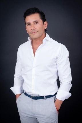 Un hombre con una camisa blanca y pantalones grises está de pie con las manos en los bolsillos.