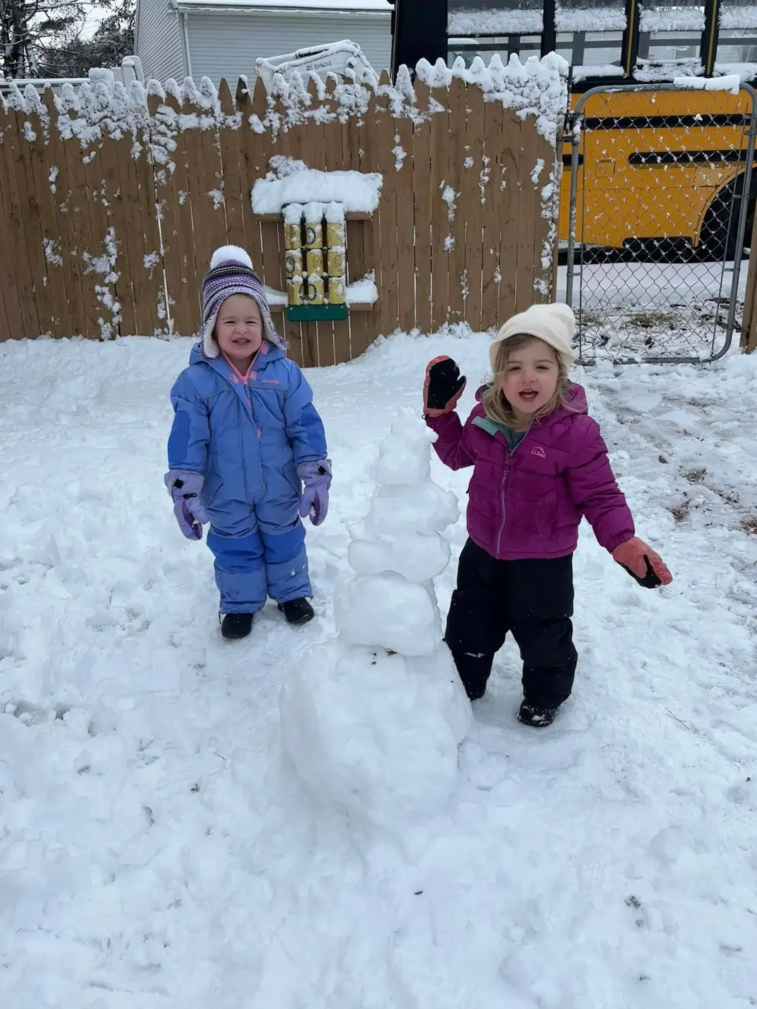 Two little girls are playing in the snow with a bird feeder in the background