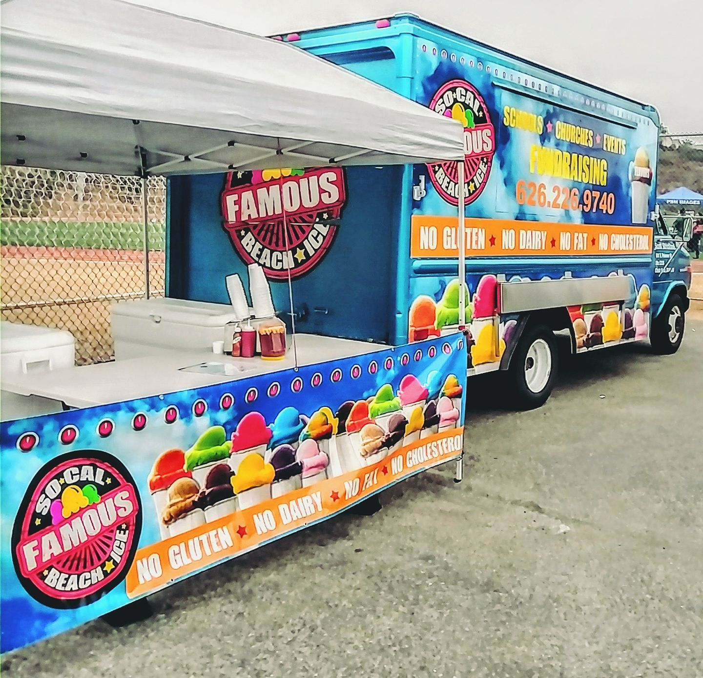 So-Cal Famous Beach Ice food truck with a branded pop-up tent, set up on a paved surface near a baseball field.