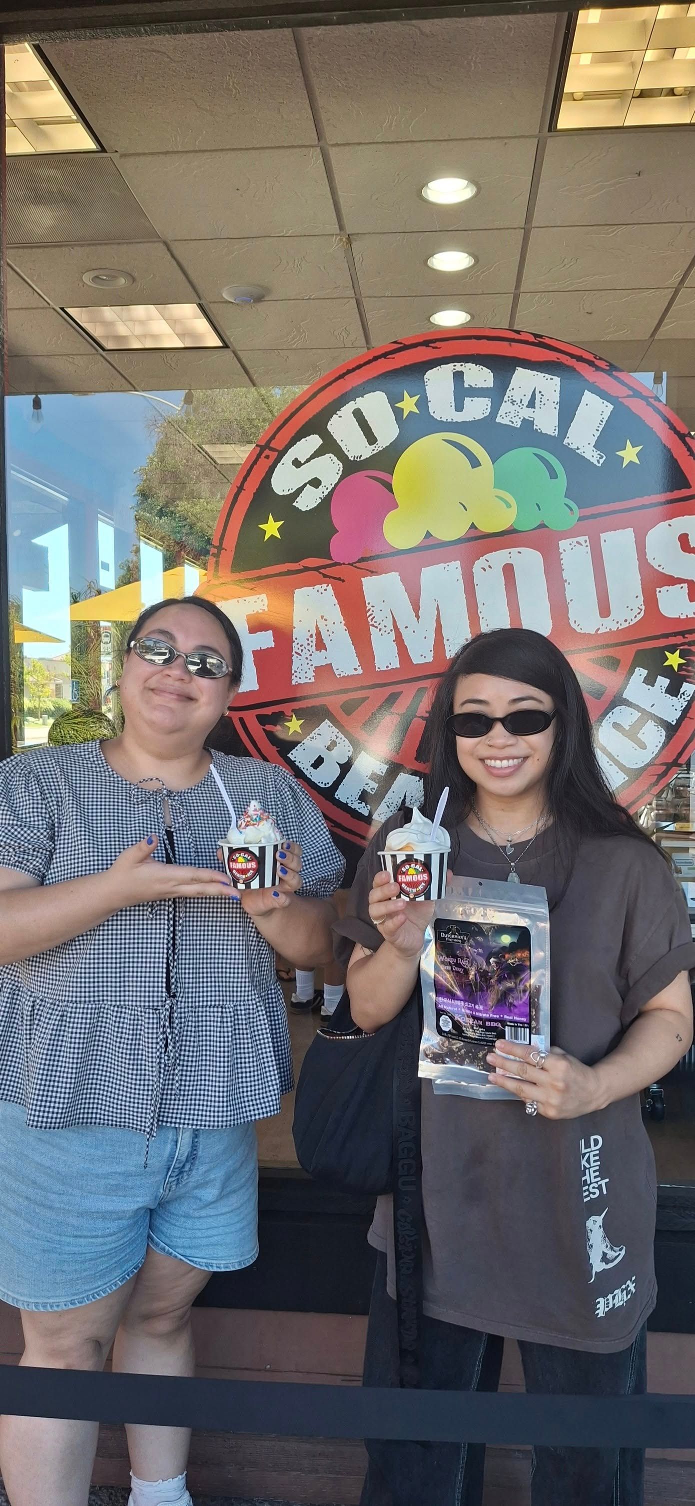 Two people stand in front of a So-Cal Famous Ice Cream window, smiling and holding small cups and bags of treats.
