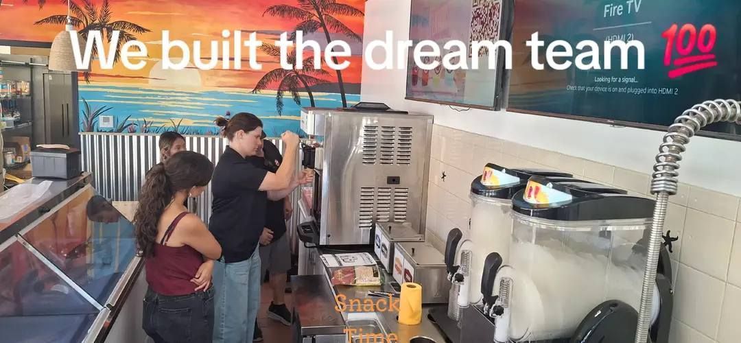 Employees work behind the counter of a snack shop decorated with a beach mural, with text overlay reading.