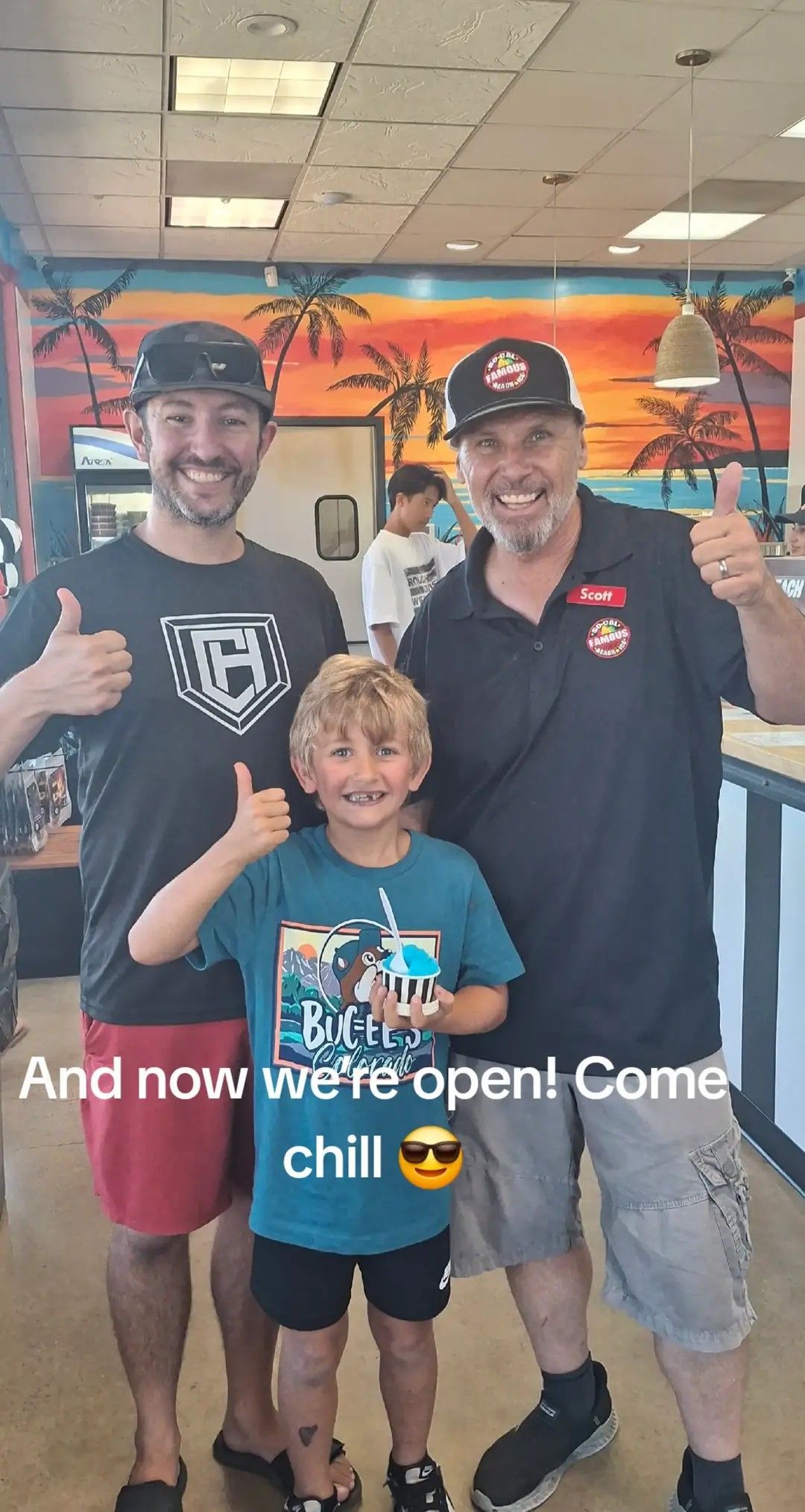 Three people smile and give a thumbs-up in a beach-themed shop with a mural behind them, celebrating an opening.
