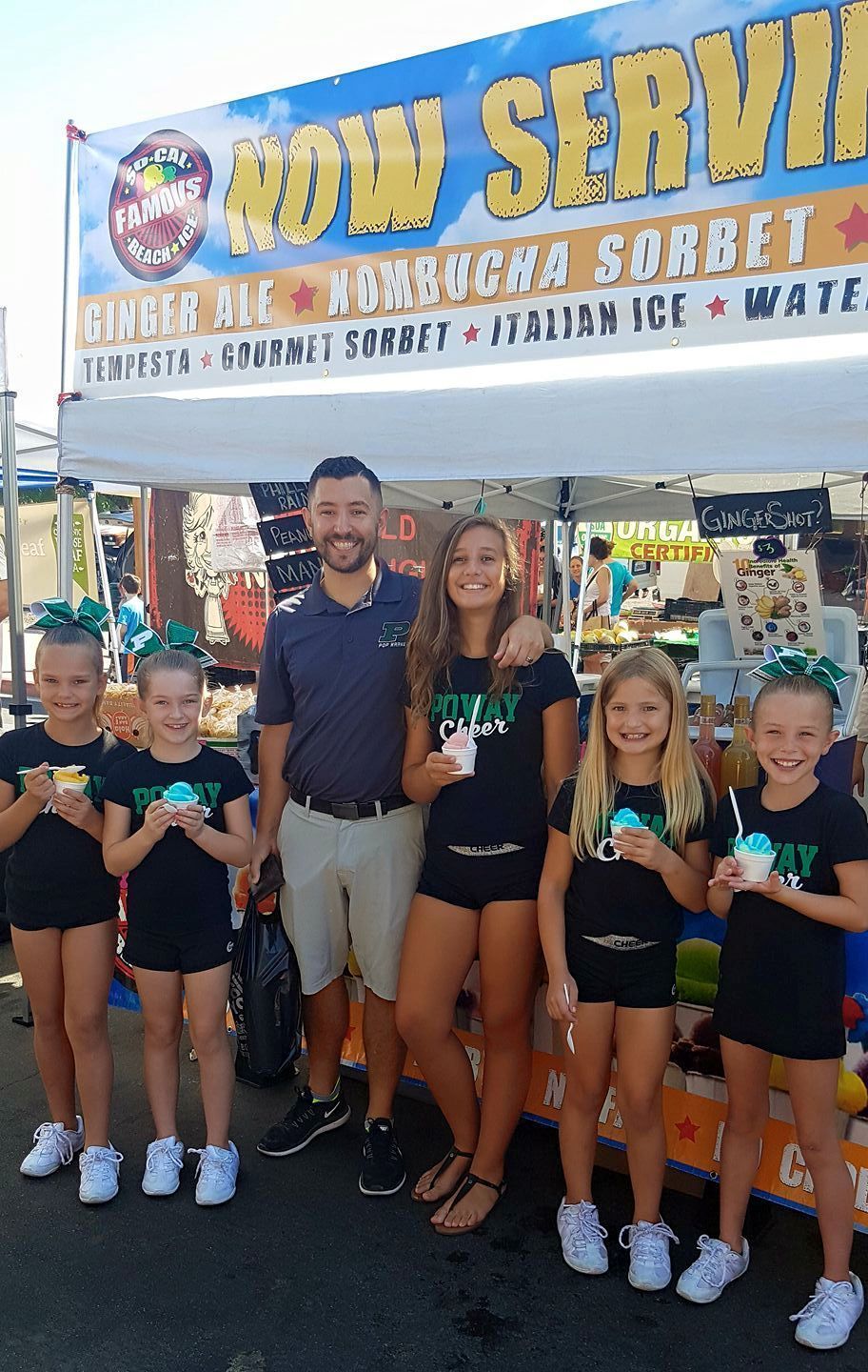 A smiling man and five girls in matching shirts stand in front of a “Now Serving” frozen treat stand holding shaved ice.
