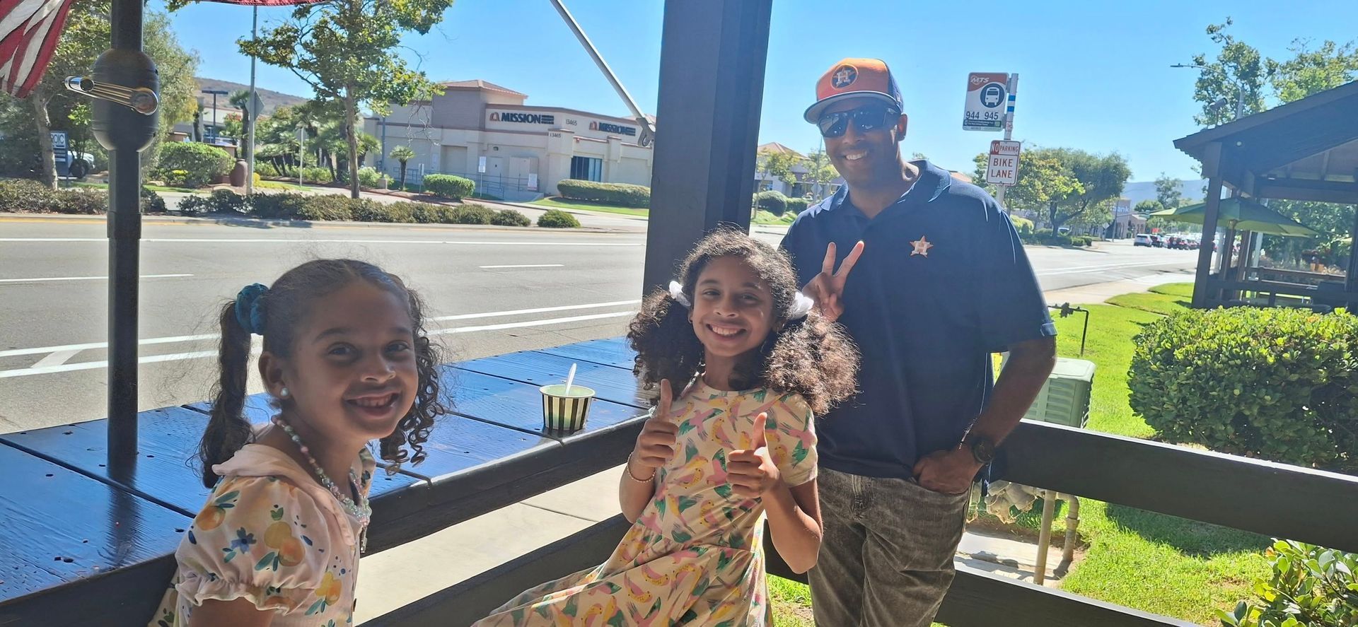 Two children and a man smiling and posing together on a sunny outdoor patio with a road and greenery in the background.