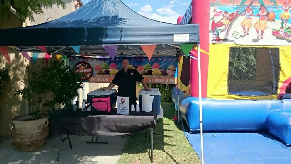 A person stands behind a table under a blue canopy at a booth, next to a large, colorful inflatable bounce house.