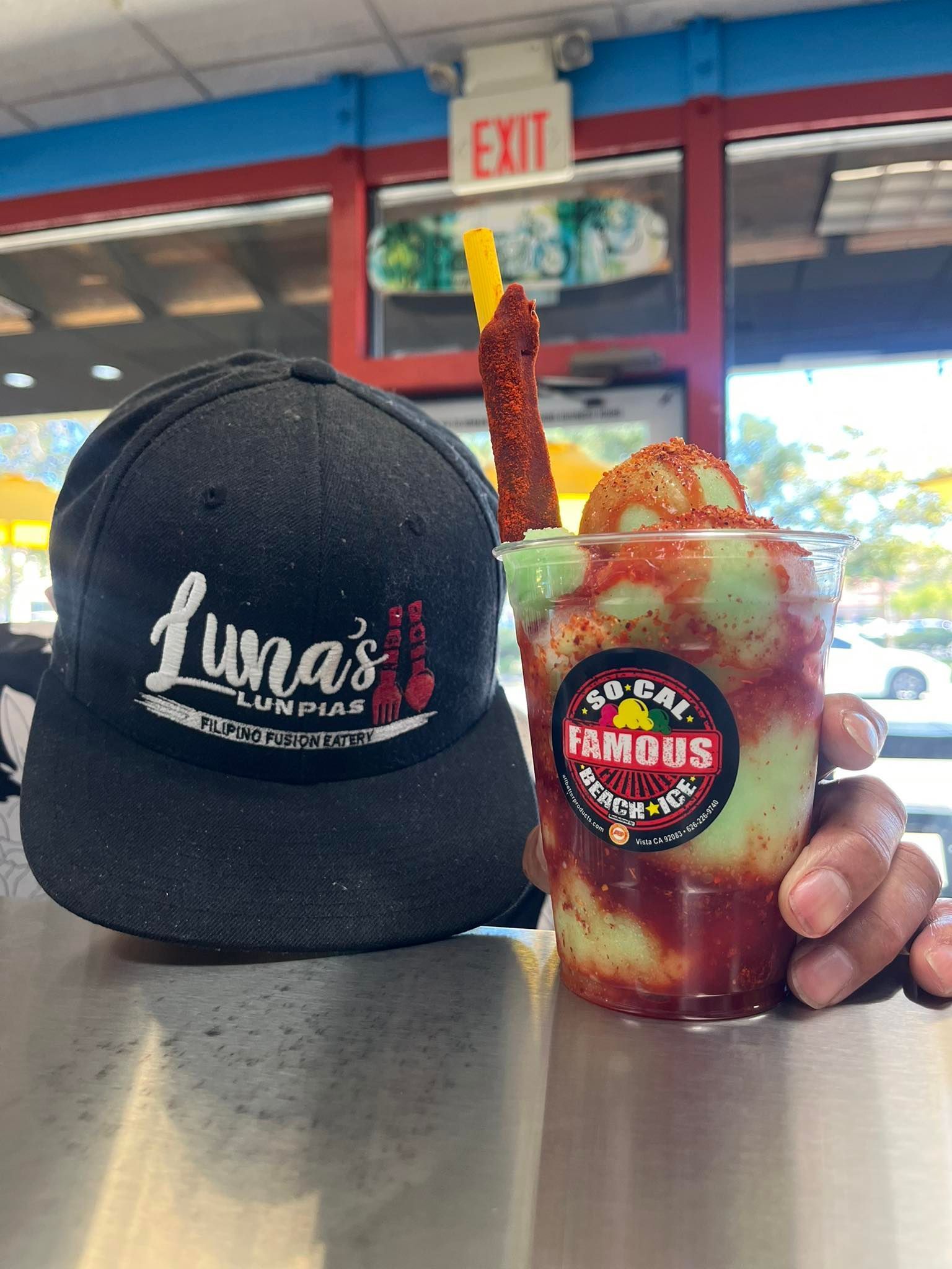 A hand holds a cup of chamoy-topped lime sorbet next to a Luna’s branded black baseball cap on a counter.