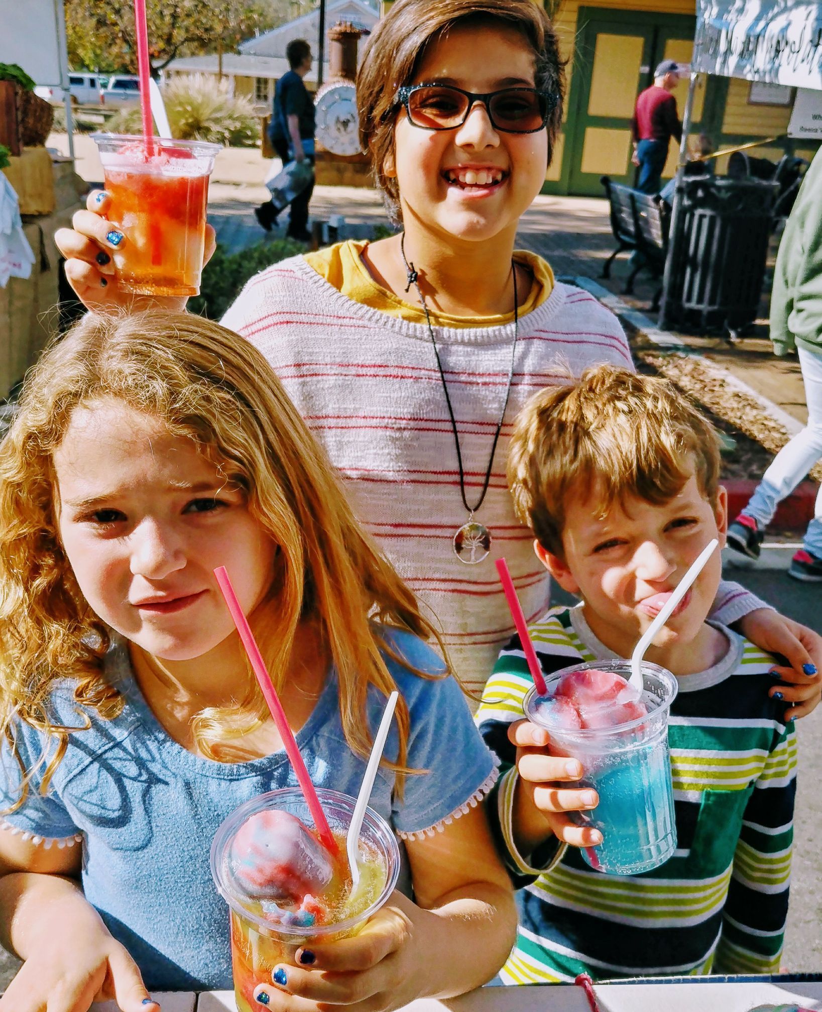 Three children smiling while holding clear plastic cups with colorful drinks and straws at an outdoor event.