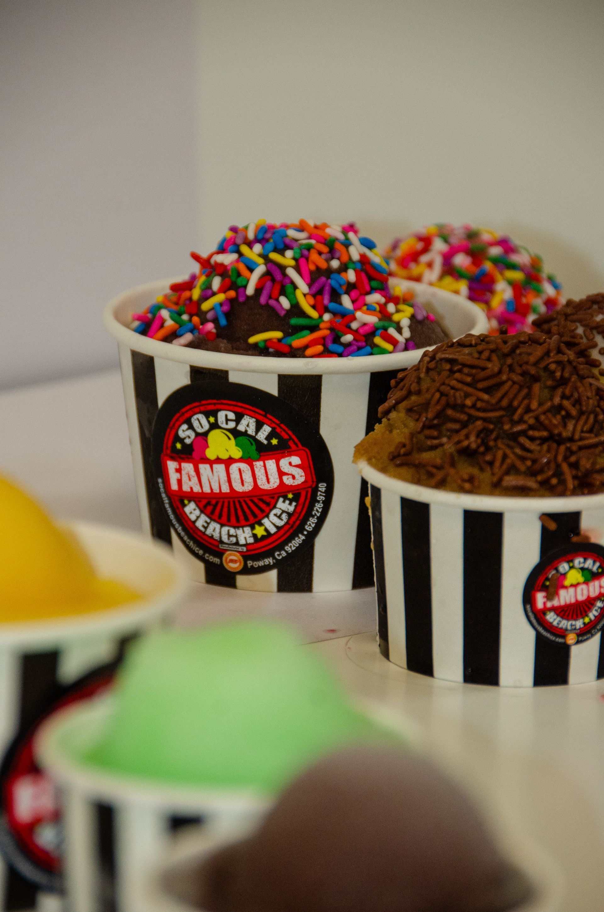 Multiple cups of colorful shaved ice and ice cream, some topped with sprinkles, featuring the So-Cal Famous Beach Ice logo.