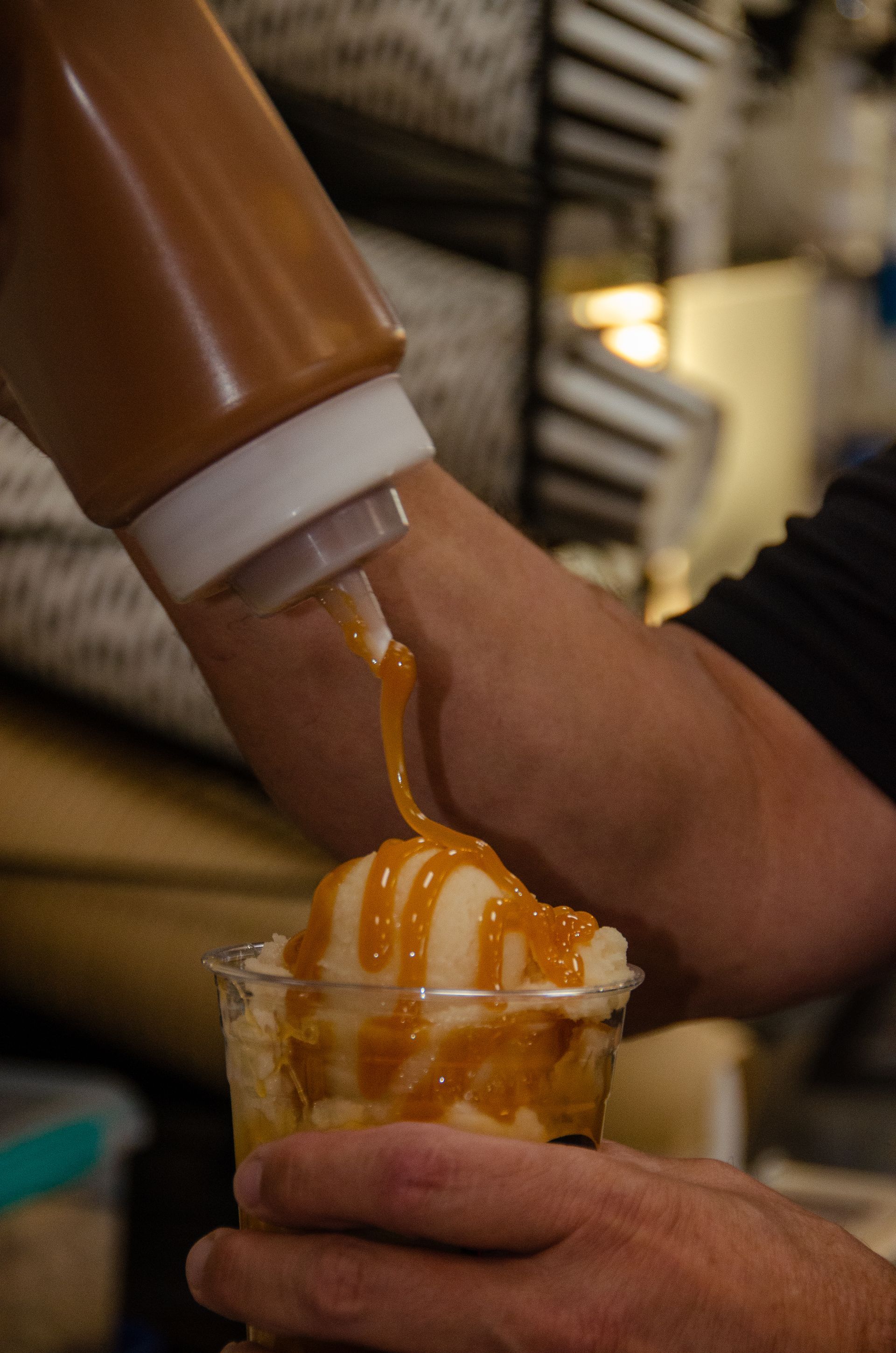 A person drizzles amber-colored caramel sauce from a squeeze bottle over a scoop of light-colored dessert in a clear cup.