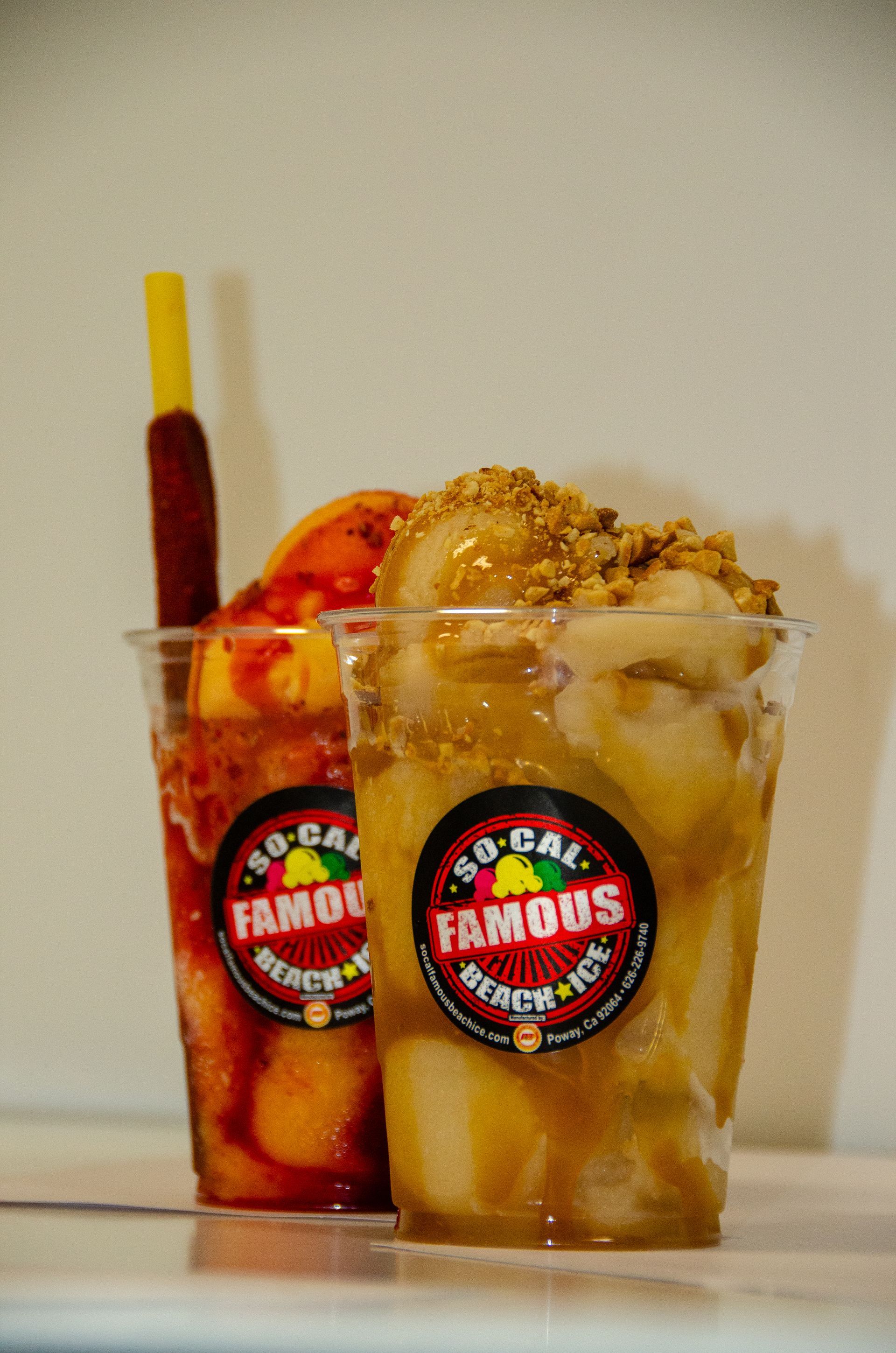 Two clear plastic cups of So-Cal Famous Beach Ice topped with fruit, sauces, nuts, and a tamarind stick.